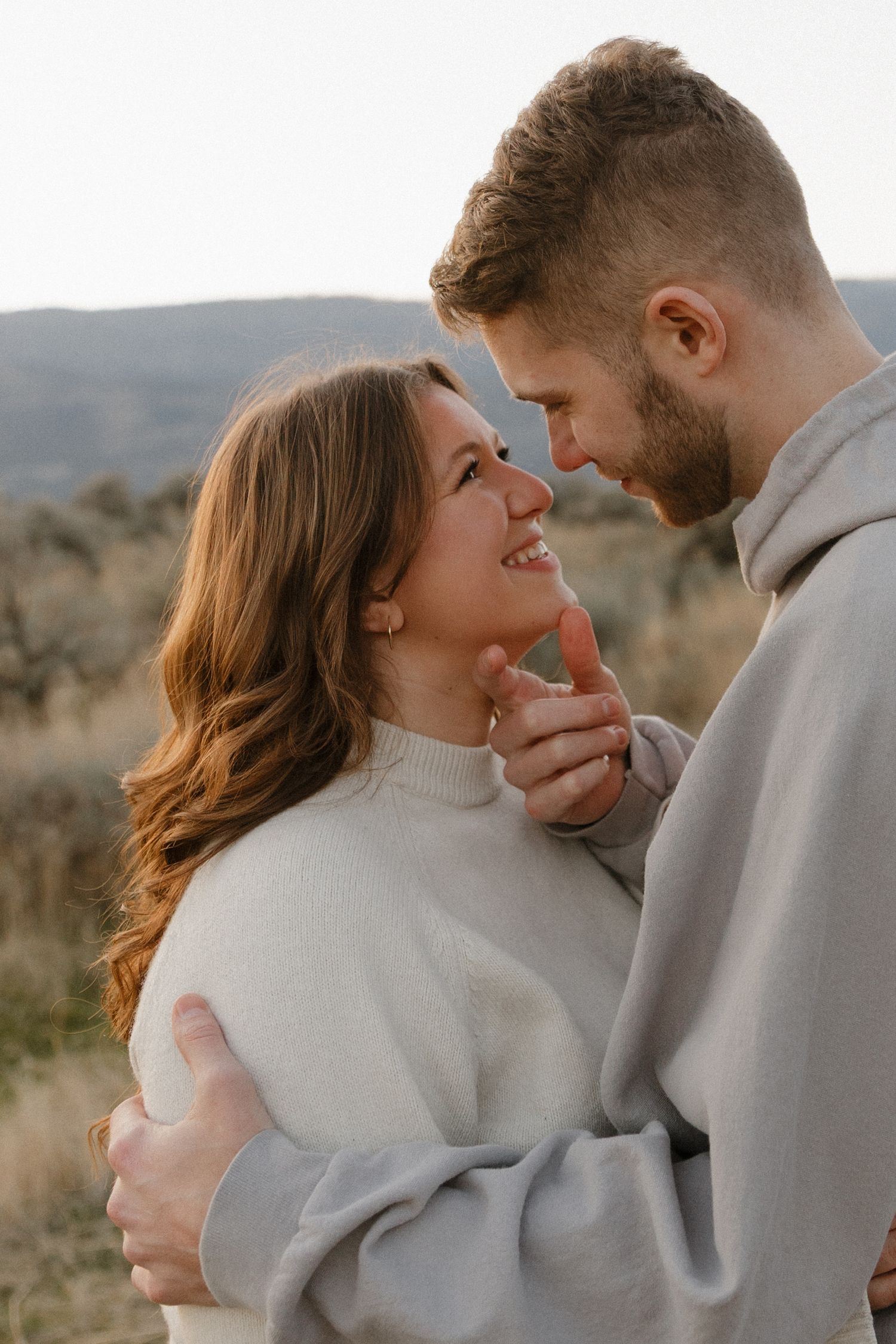 Couple embracing and smiling at each other in a golden field during a romantic outdoor engagement session.