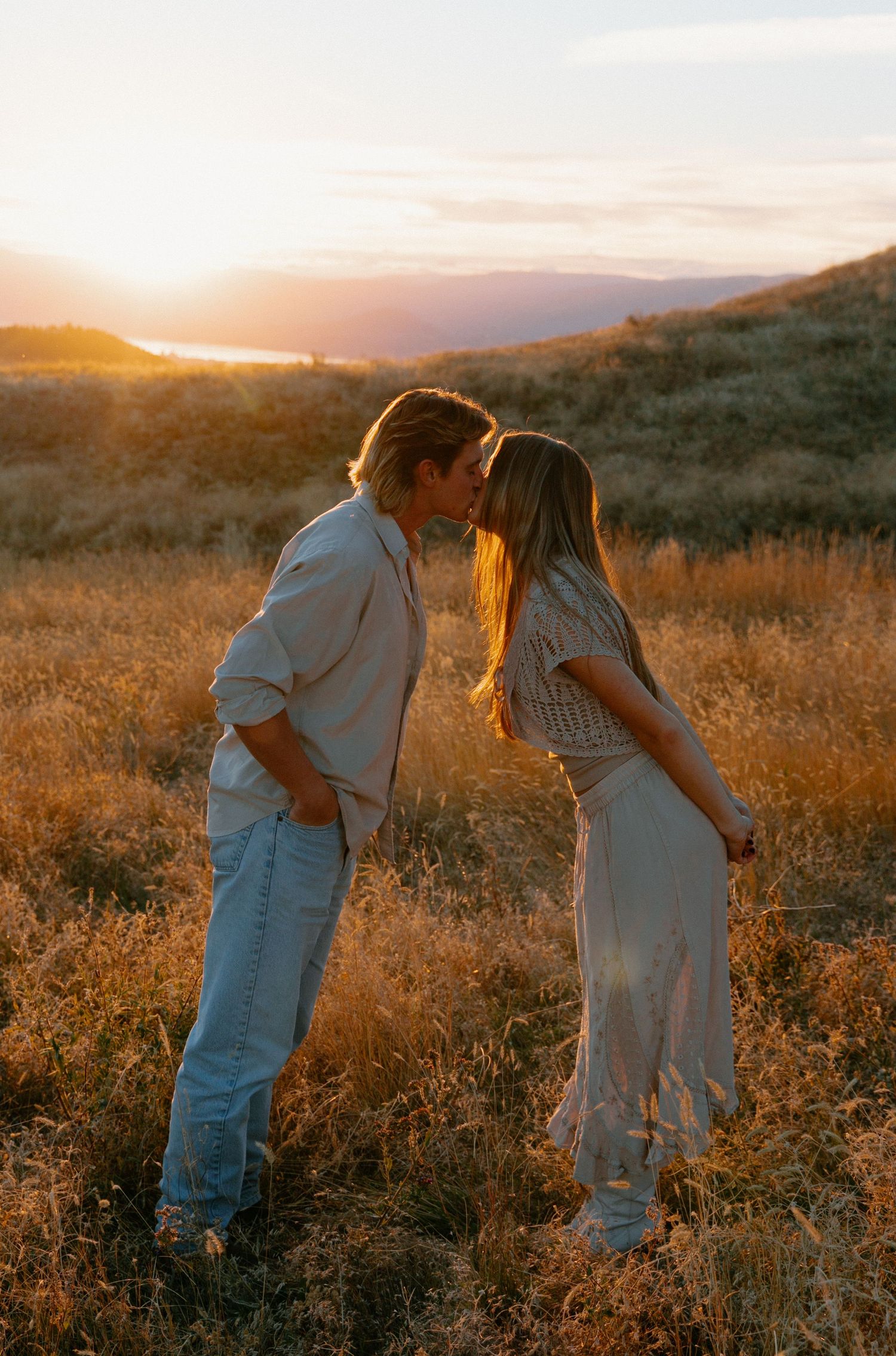 Romantic couple sharing a kiss in golden sunset field with mountain backdrop.