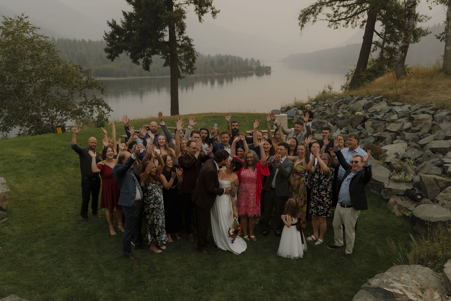 Large wedding party group photo outdoors by a lake raising glasses in celebration.