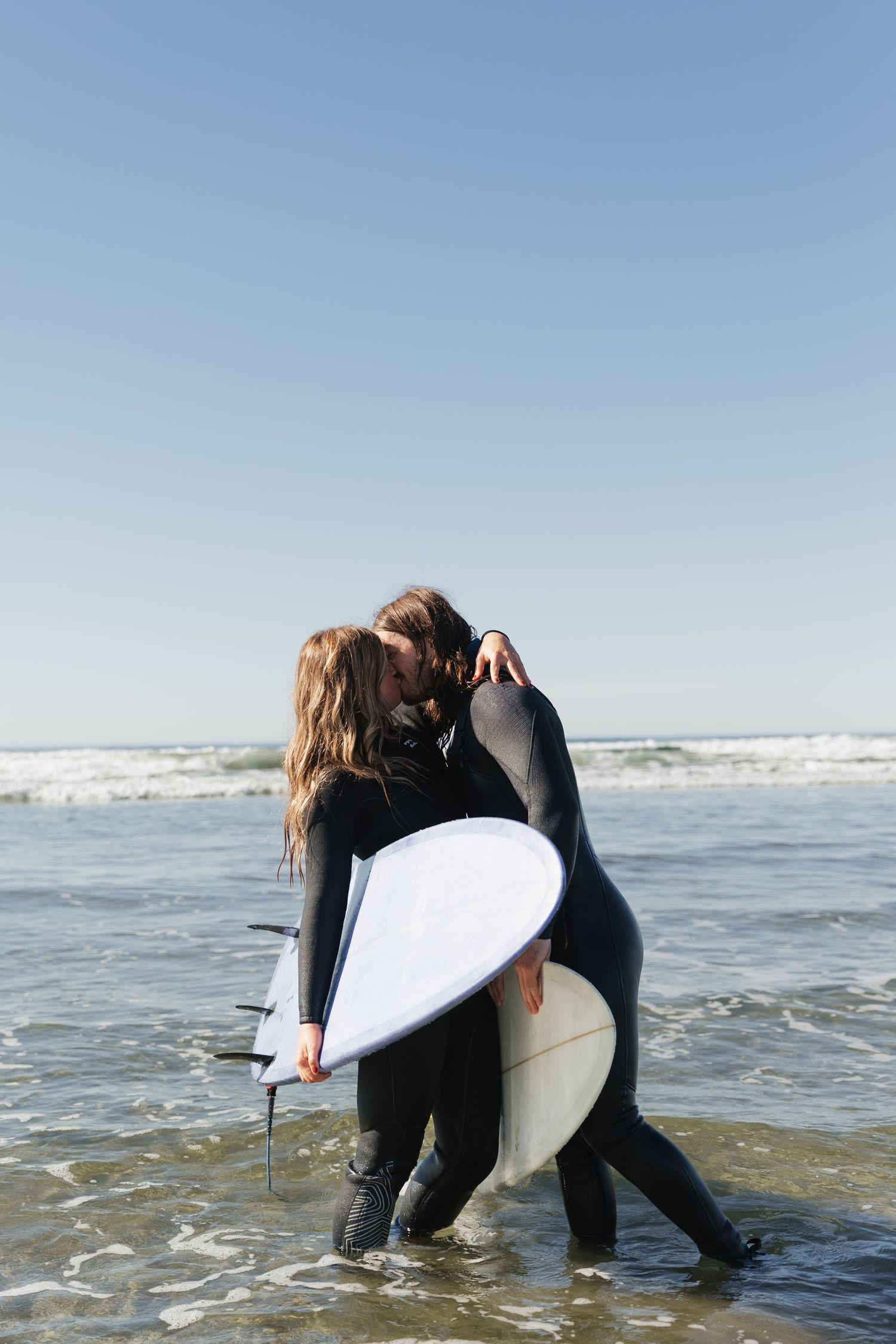Couple in black wetsuits kissing while holding a surfboard in ocean waves on a sunny day.