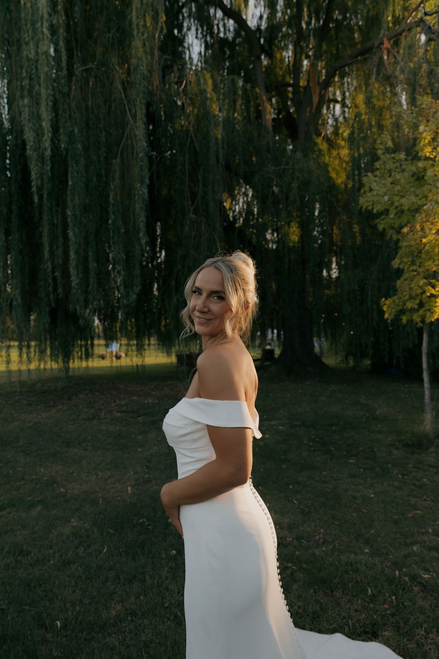 Bride in off-shoulder white gown smiles over shoulder beneath a weeping willow at sunset.