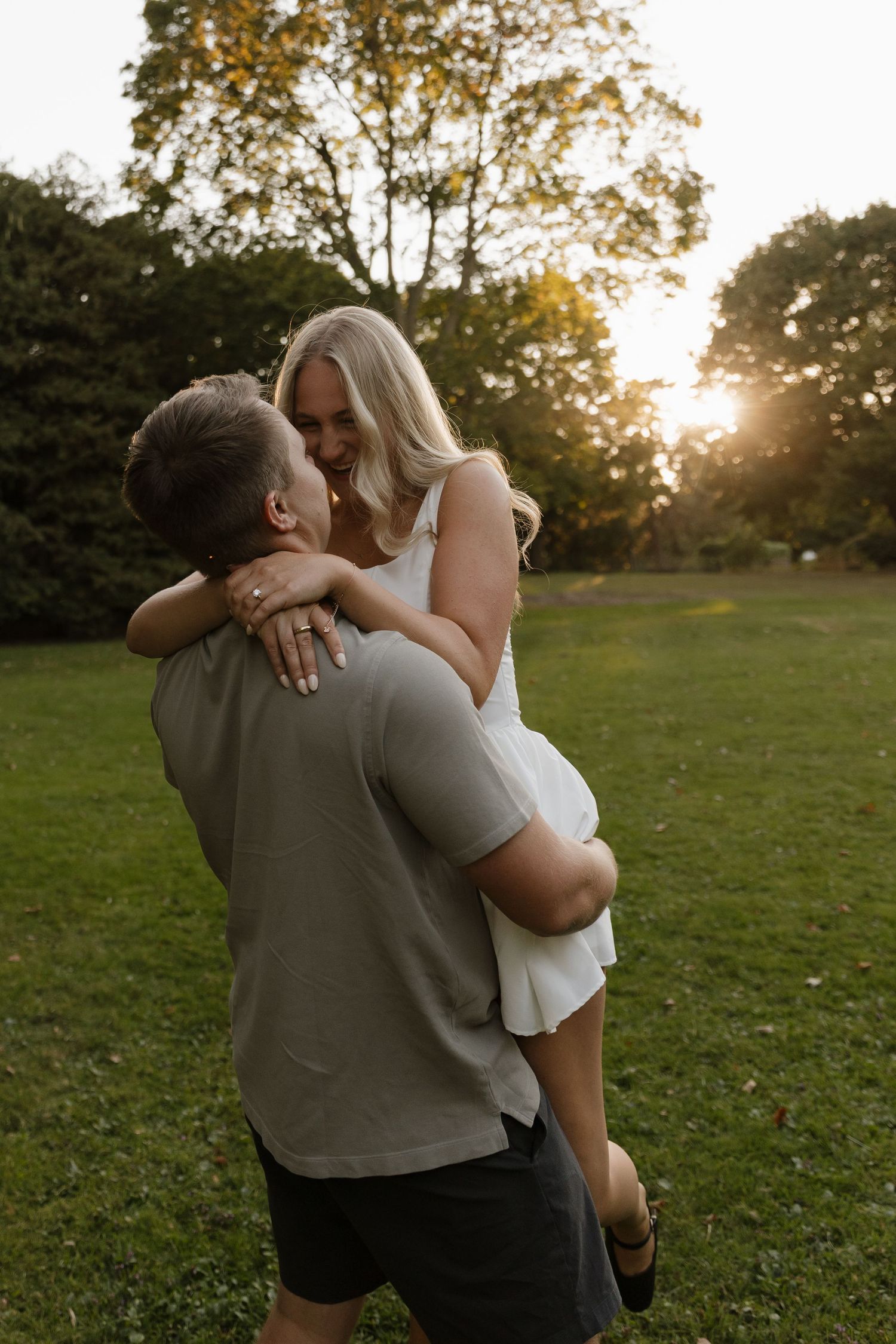 Man lifts smiling blonde woman in white dress during golden hour in a lush green park.
