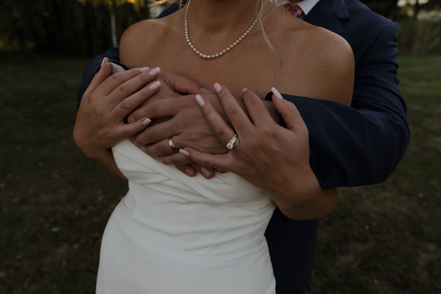 Close-up of bride and groom showcasing wedding rings, brides pearl necklace visible against white gown.