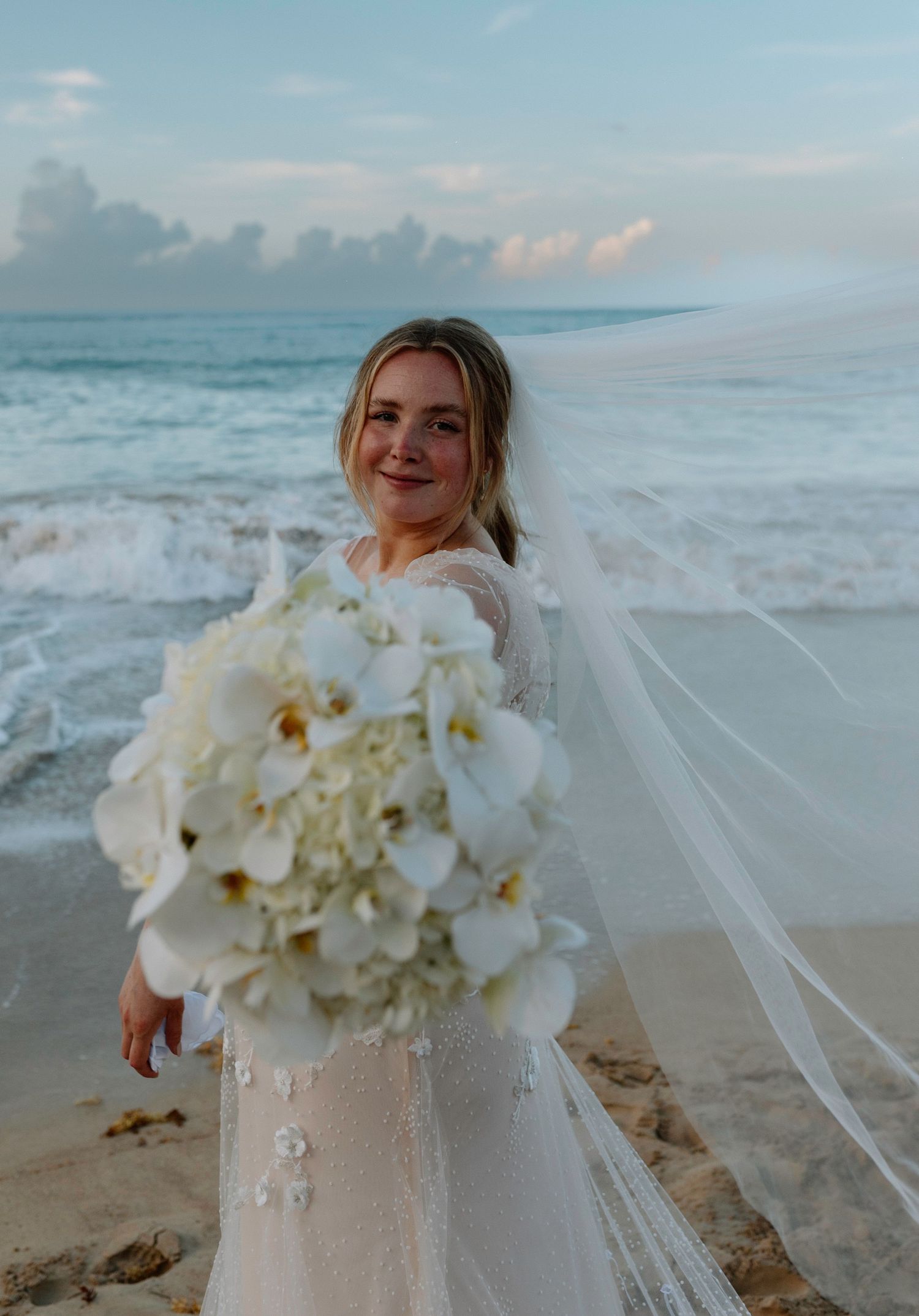 Bride in lace gown holding white orchid bouquet with flowing veil on tropical beach at sunset.