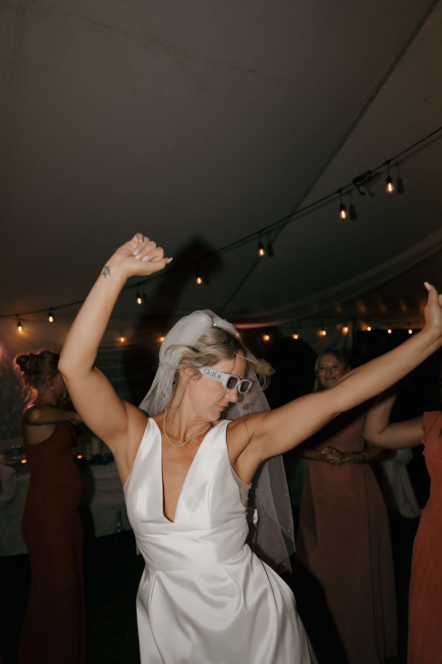 Bride in white gown and sunglasses dancing with arms raised at wedding reception.