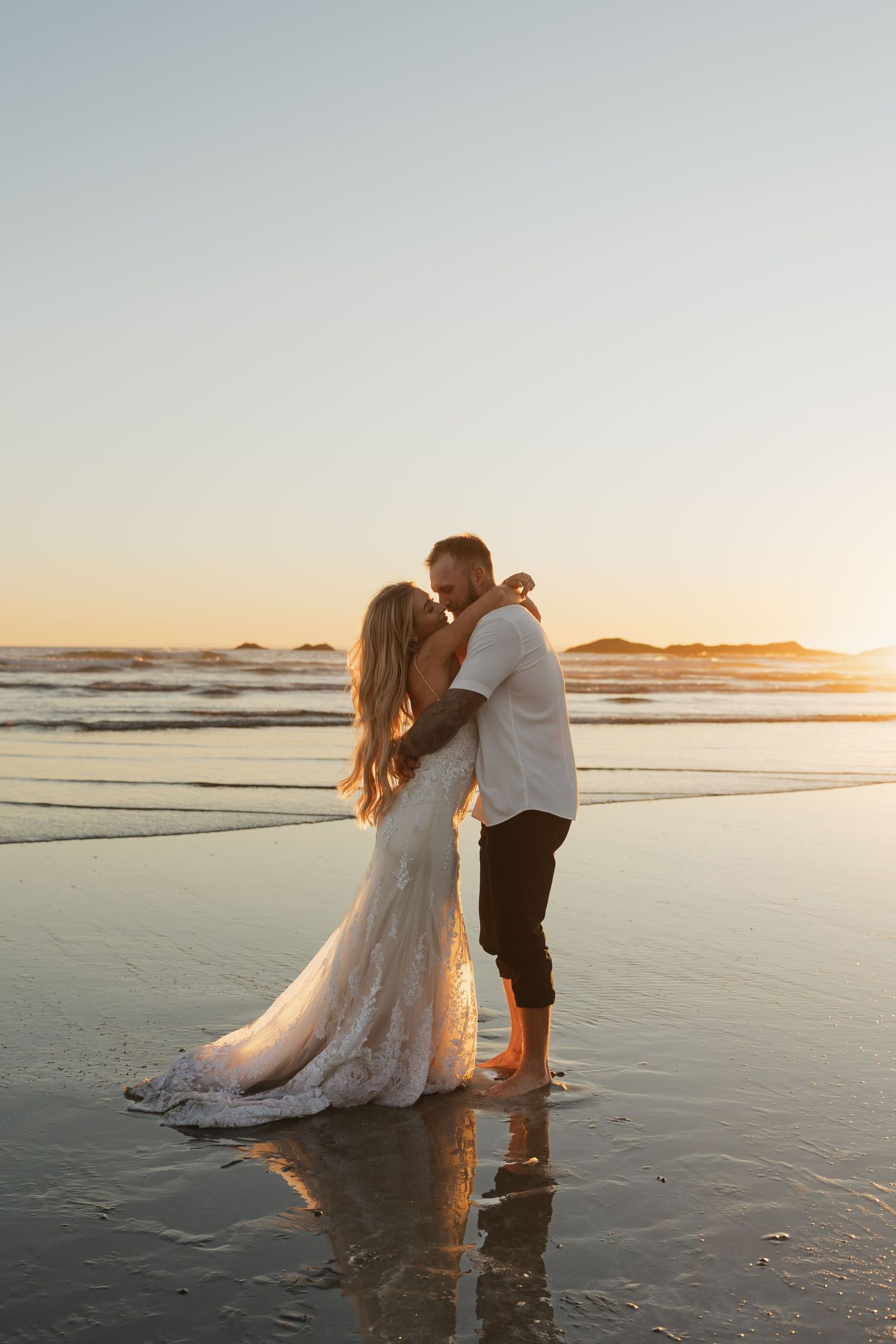 Newlyweds share a kiss standing in shallow beach water at sunset, bride in flowing lace gown.