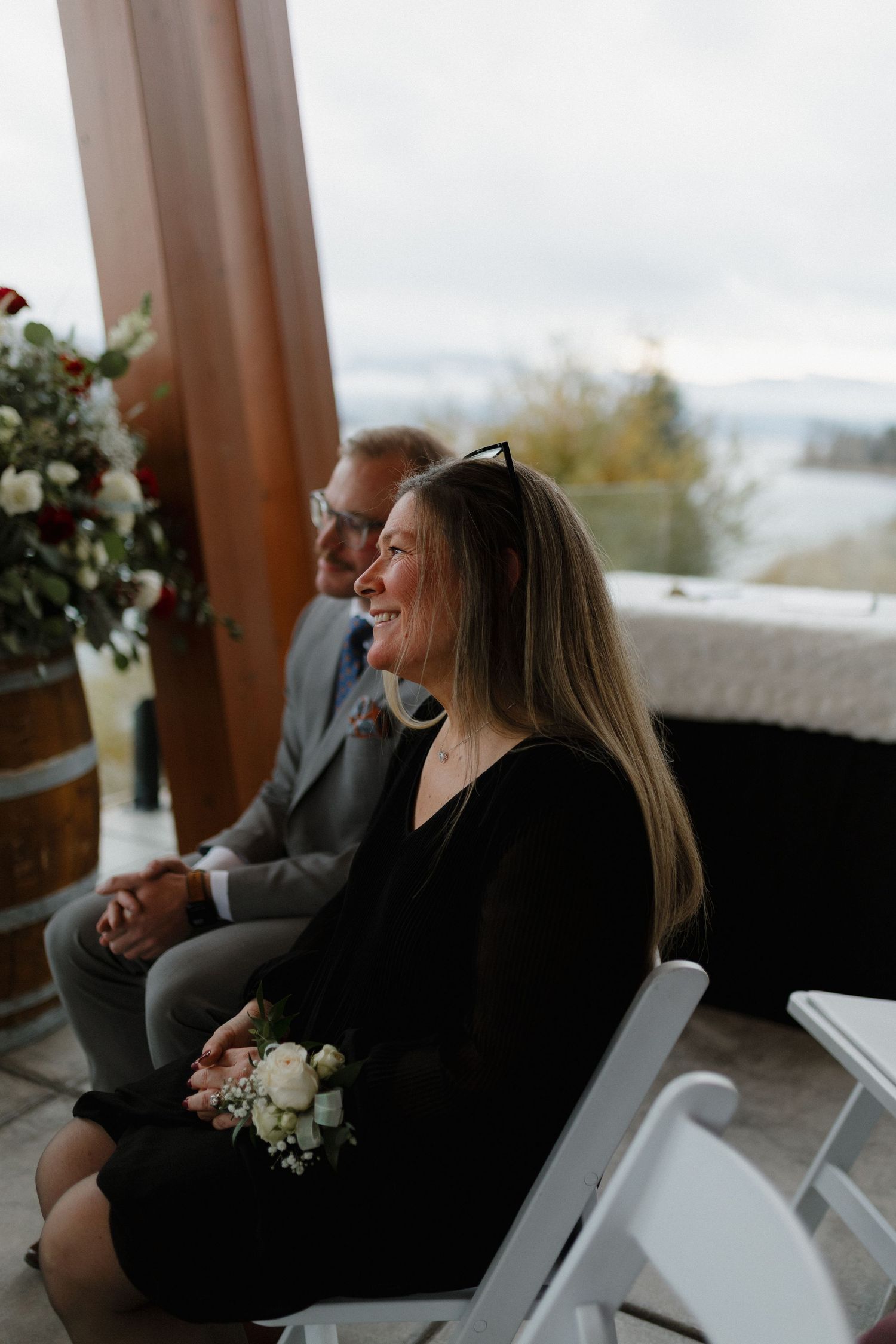 Smiling female wedding guest in black dress holding white flower corsage seated at wedding ceremony.