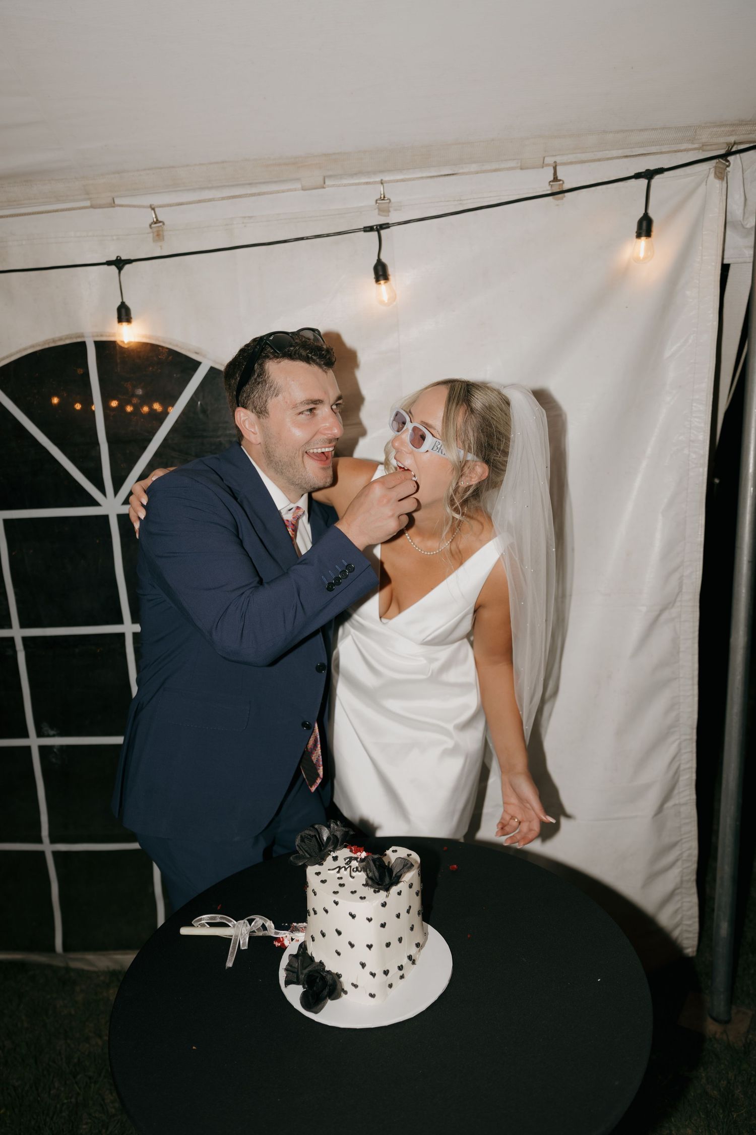 Groom feeding bride wedding cake at reception, both smiling under string lights.