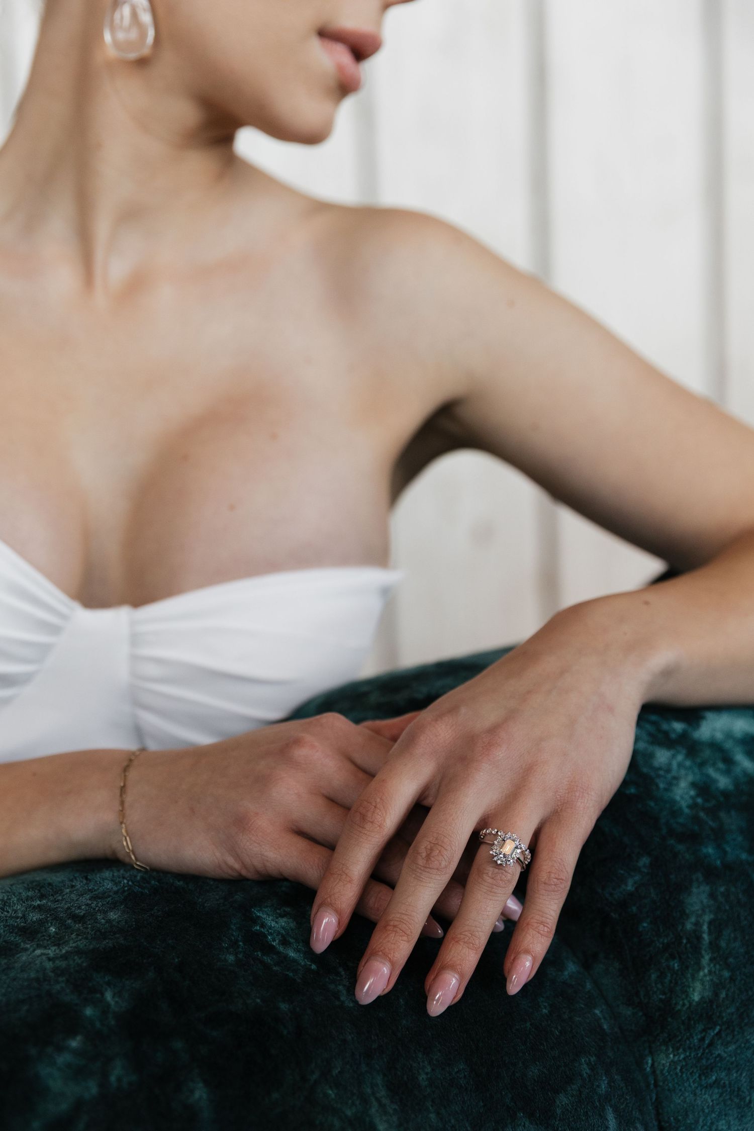 Close-up of bride in strapless white wedding dress showcasing diamond ring and gold bracelet on velvet sofa.
