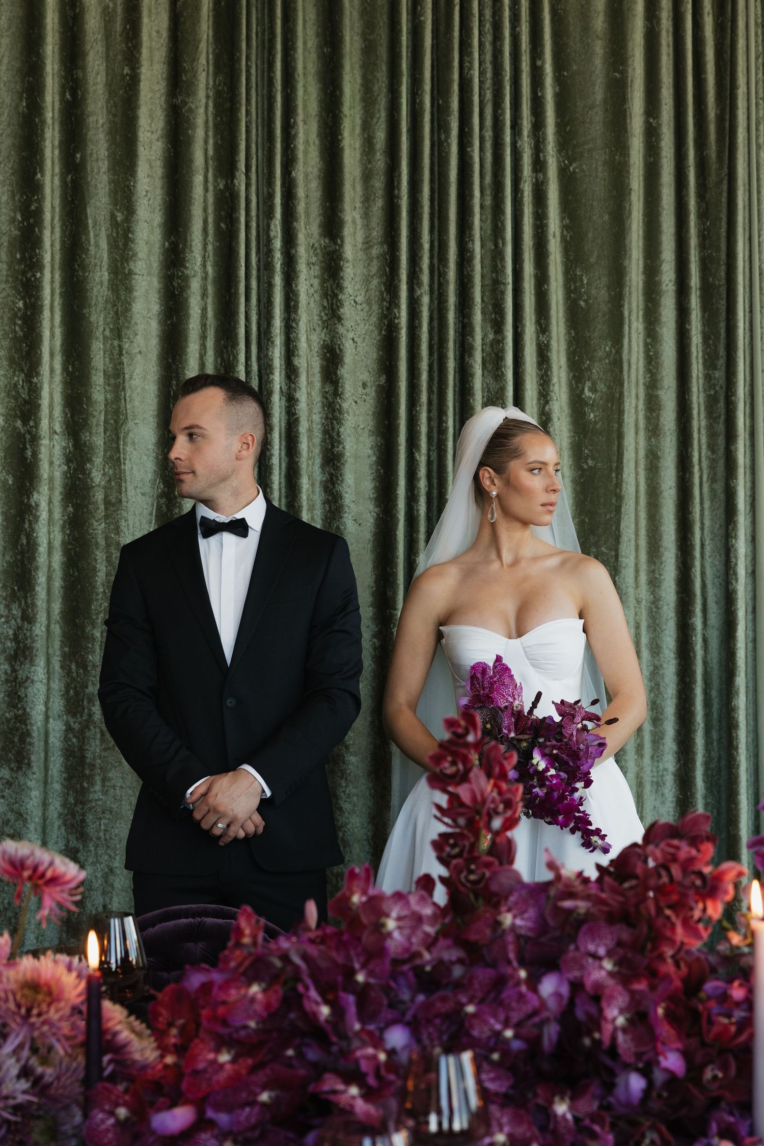 Bride holding purple orchid bouquet and groom in black tuxedo standing before green velvet curtain at ceremony.