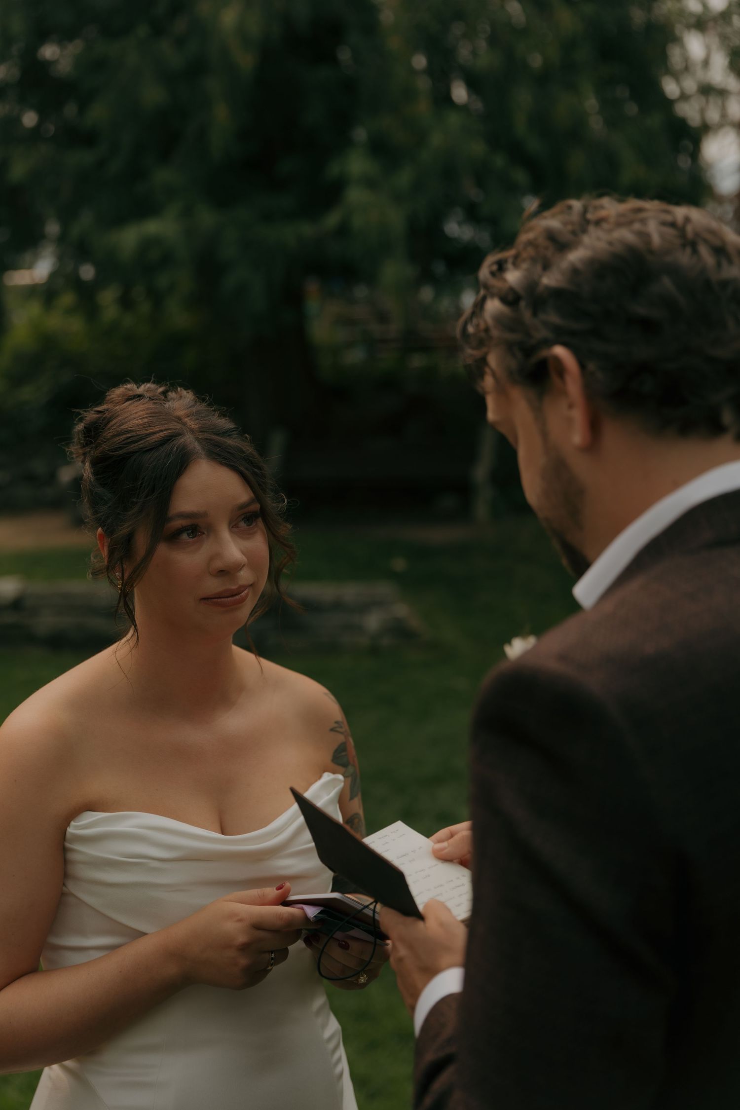 Bride in strapless white gown reading wedding vows to groom during an intimate outdoor ceremony.