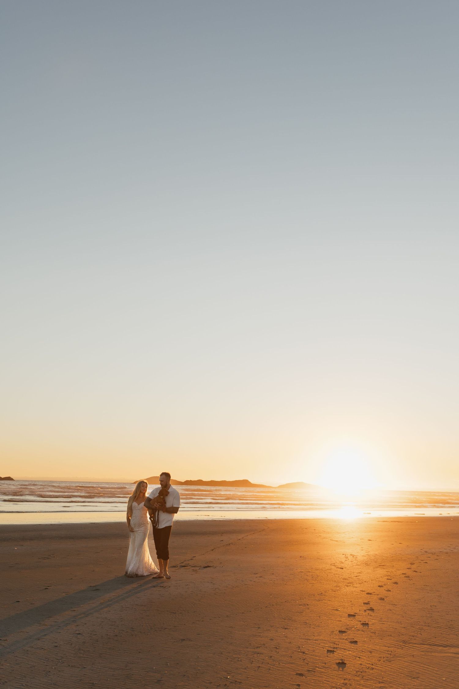 Newlyweds standing together on a sandy beach during a golden sunset with ocean waves behind them.