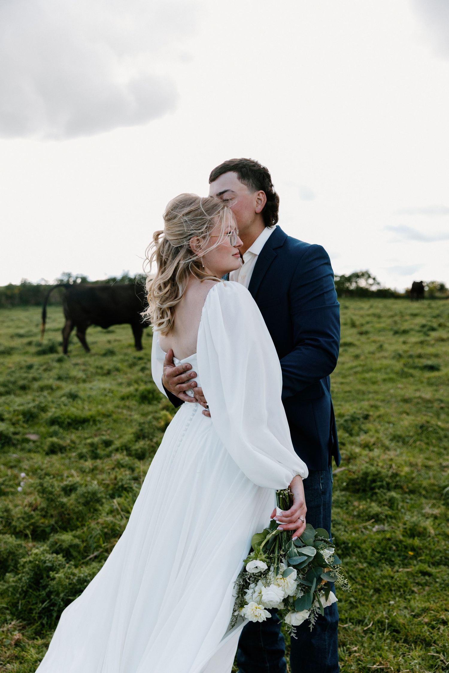 Groom tenderly kissing bride while she holds a white bouquet in a lush green pasture.
