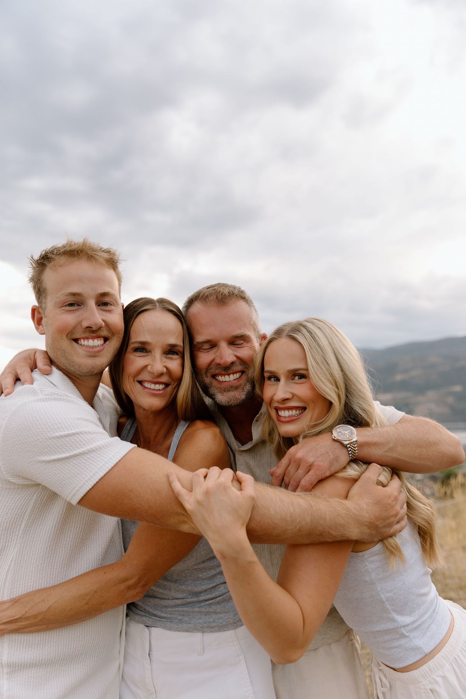 Four smiling adults hugging together outdoors with a cloudy mountain landscape in the background.