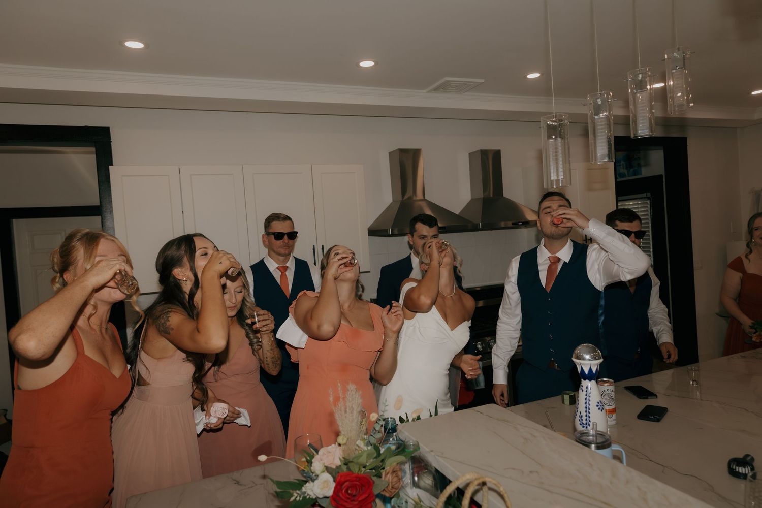 Excited wedding party does shots together in modern white kitchen during lively wedding reception celebration.