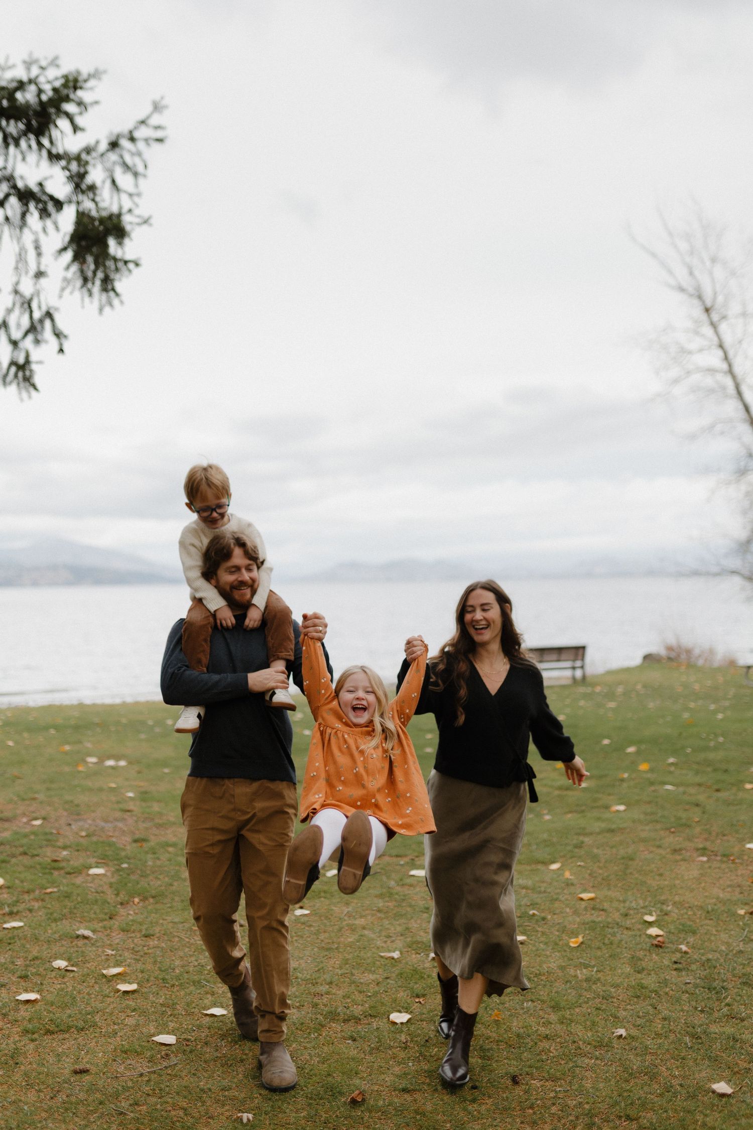Joyful family of four playing outdoors near a lake on a cloudy fall day.