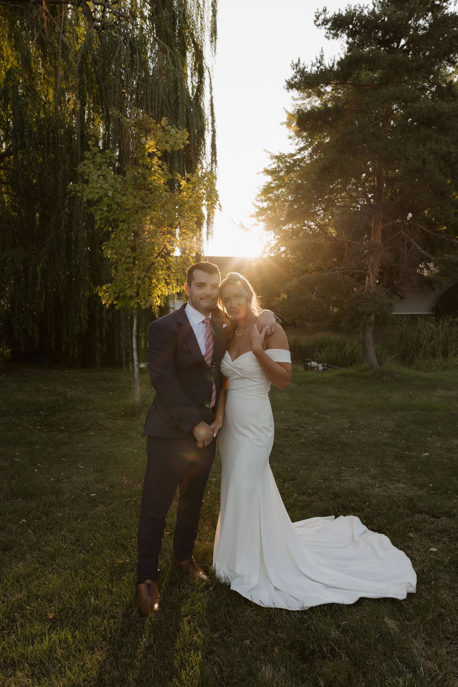 Bride and groom embrace in golden sunset light surrounded by weeping willow and tall trees.
