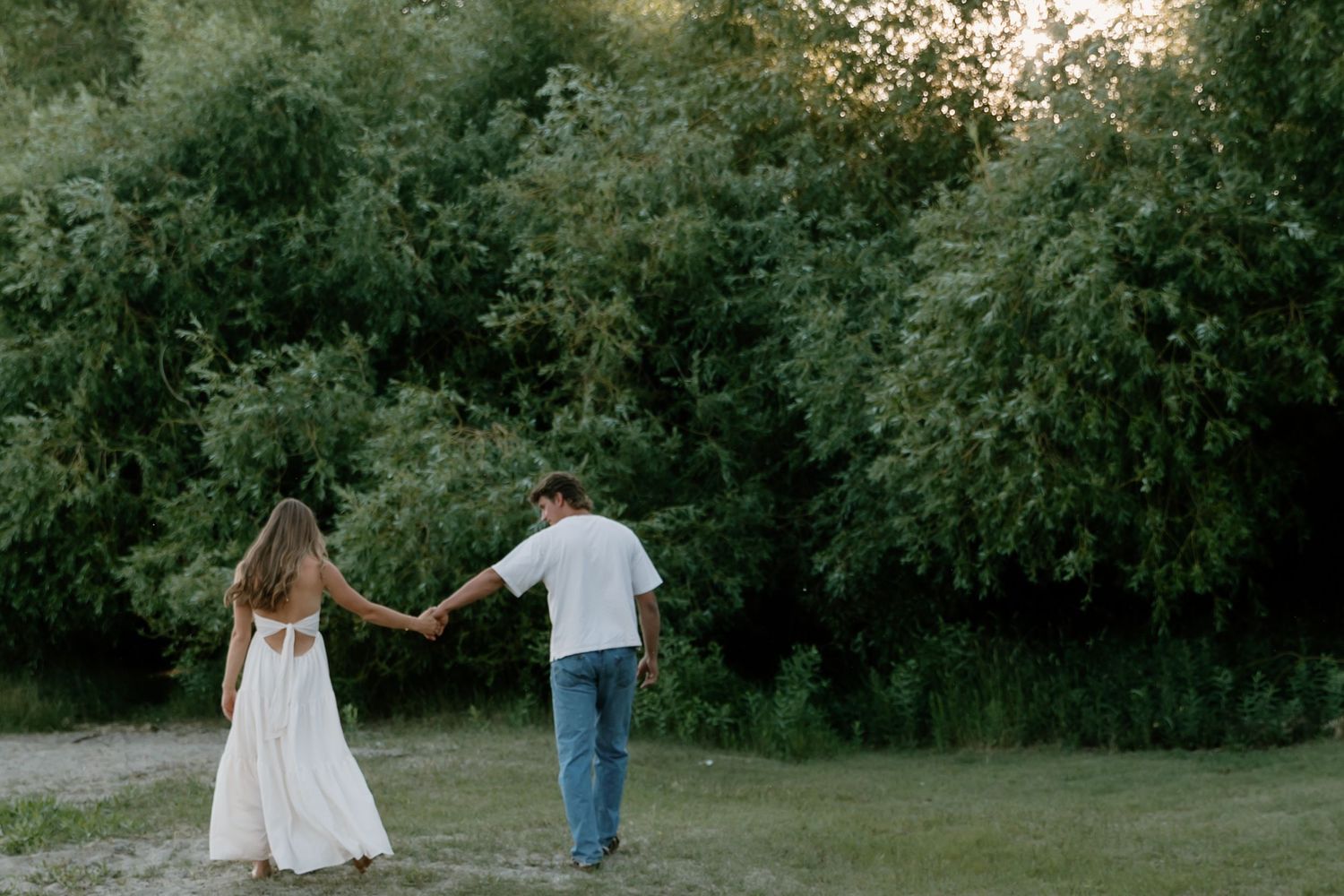 Couple walks hand-in-hand through a serene green meadow at dusk, woman in elegant white dress.