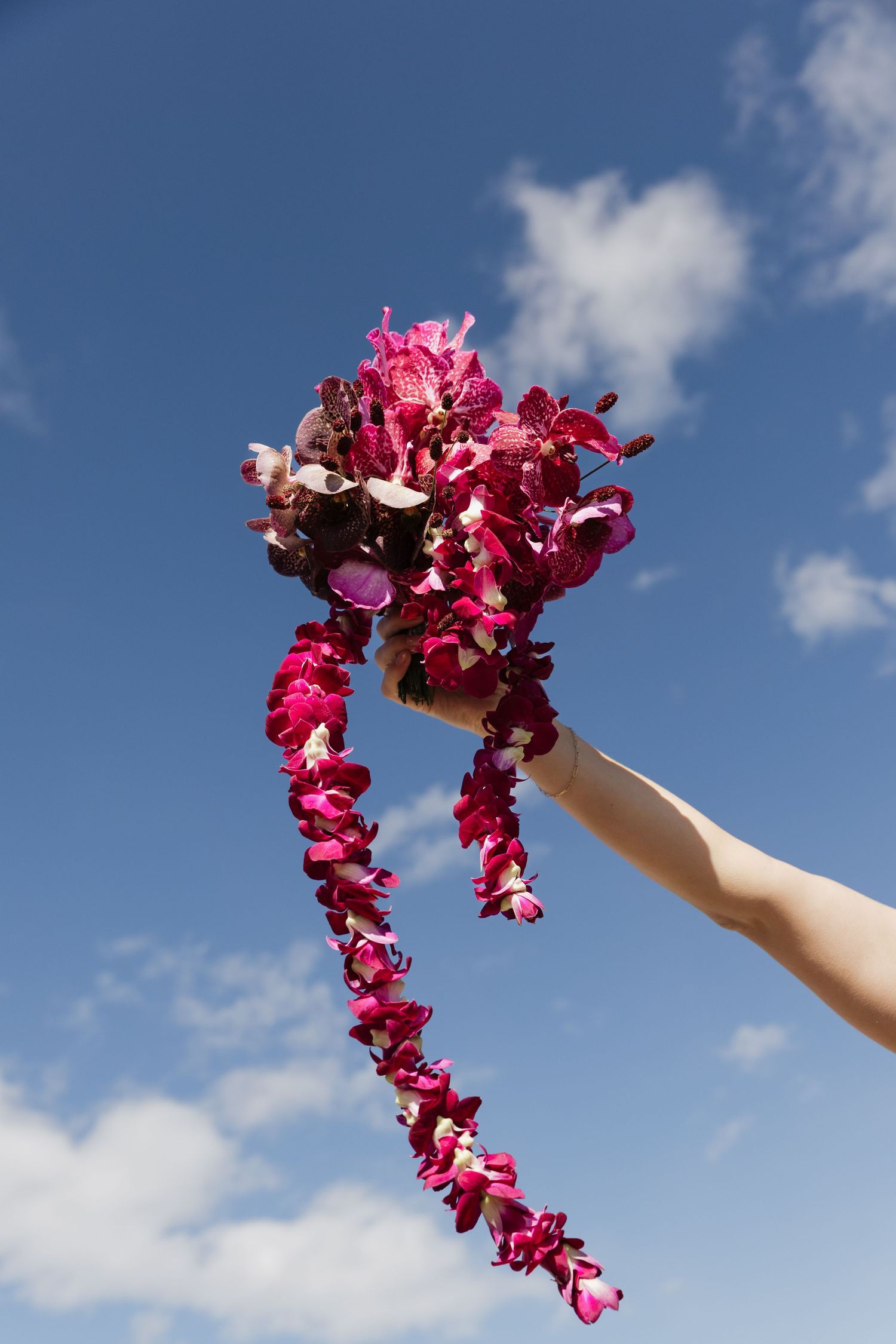 Hand raised holding a cascading pink and magenta orchid bridal bouquet against a bright blue sky.