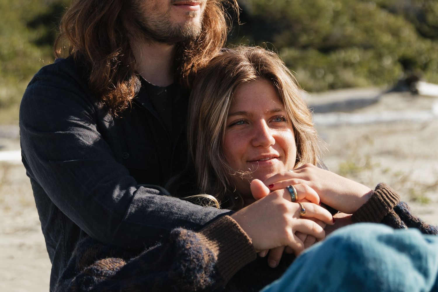Couple cuddling outdoors, woman smiling warmly while man wraps arms around her in a cozy natural setting.