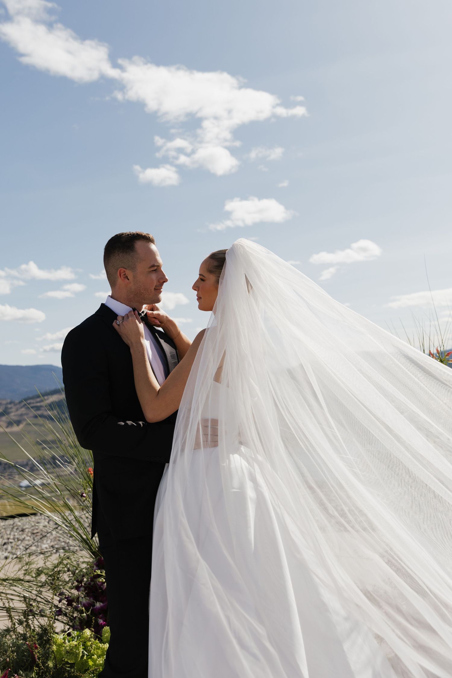 Bride adjusts grooms bow tie as her veil flows in the wind against a blue sky backdrop.