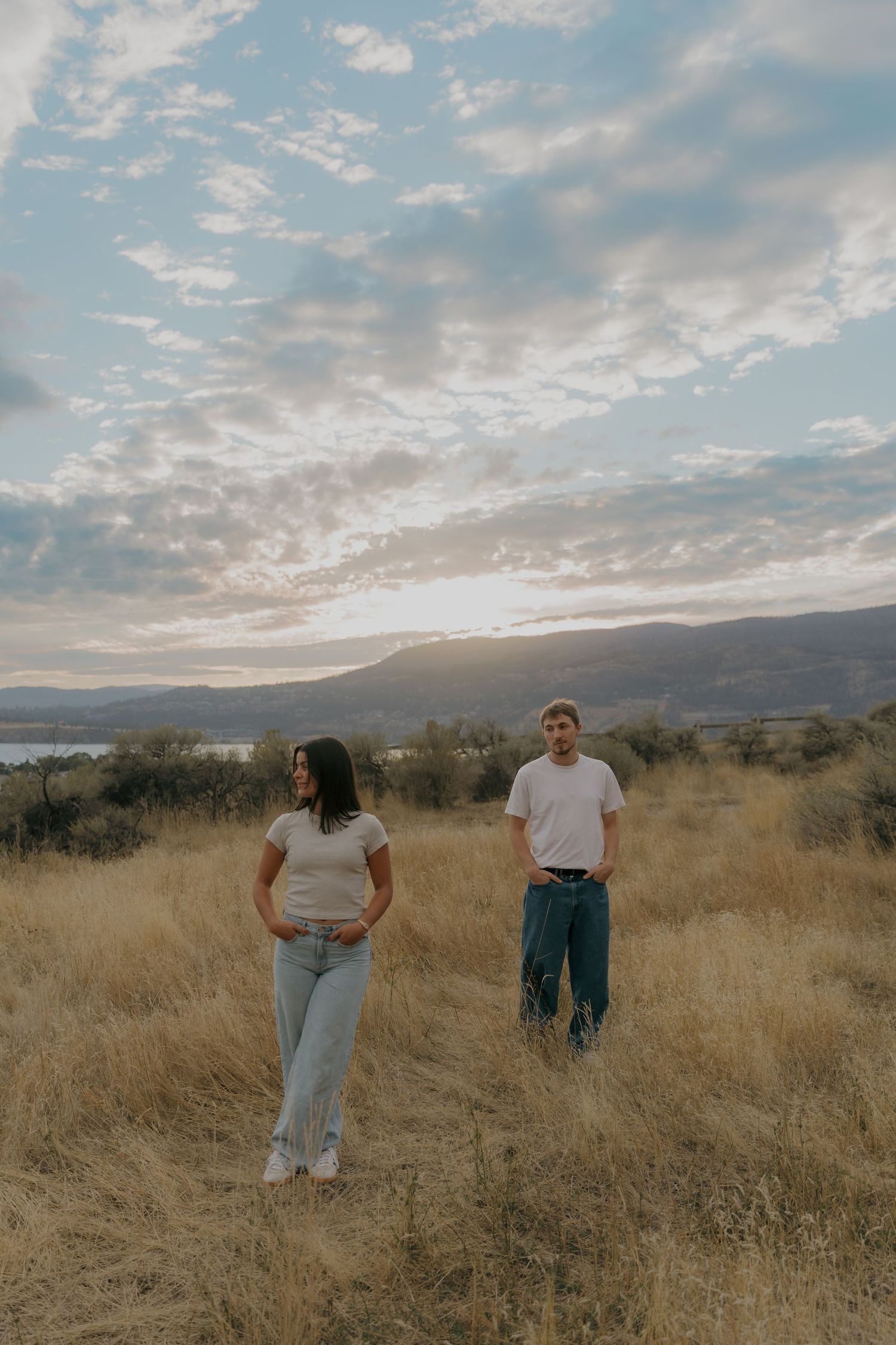 Couple stands apart in golden dry grass field at sunset with mountain lake backdrop.