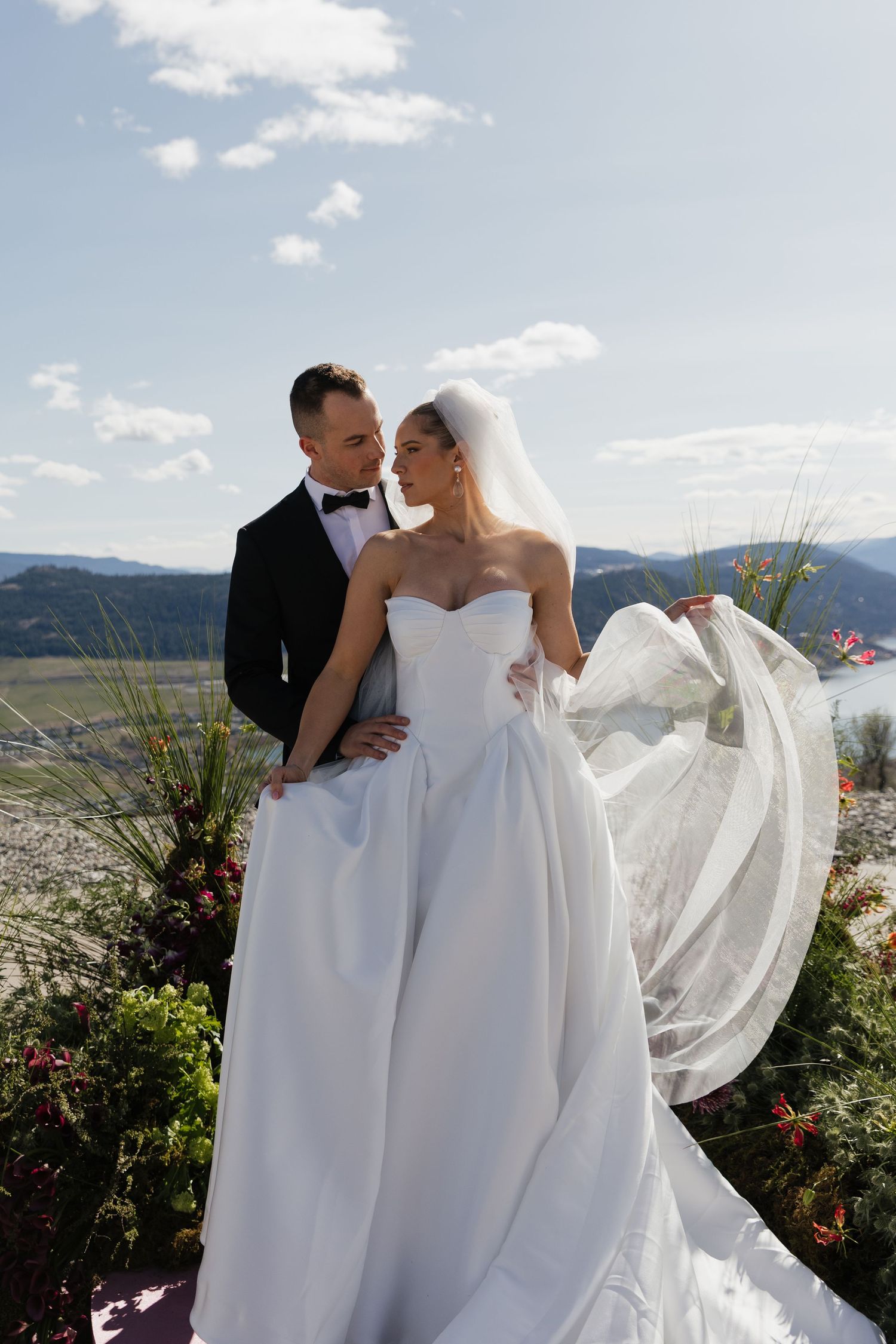 Bride in flowing white gown with veil and groom in black tuxedo posing at outdoor venue with mountain views.