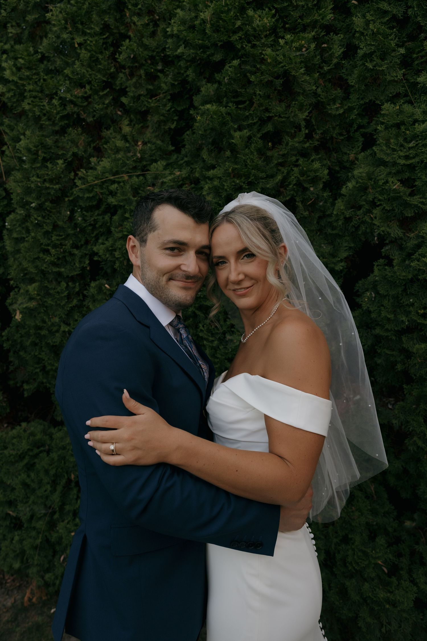 Smiling bride and groom embrace warmly before lush green evergreen hedge, bride in off-shoulder white gown with veil.