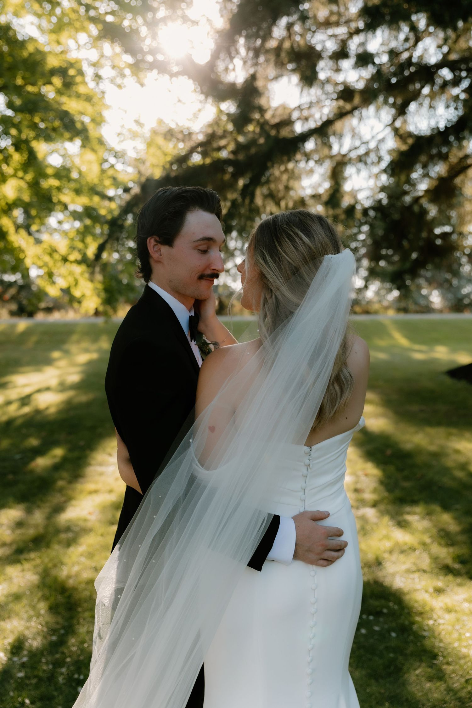 Bride and groom embrace in sunlit garden, bride's veil flowing as they lean in for a kiss.