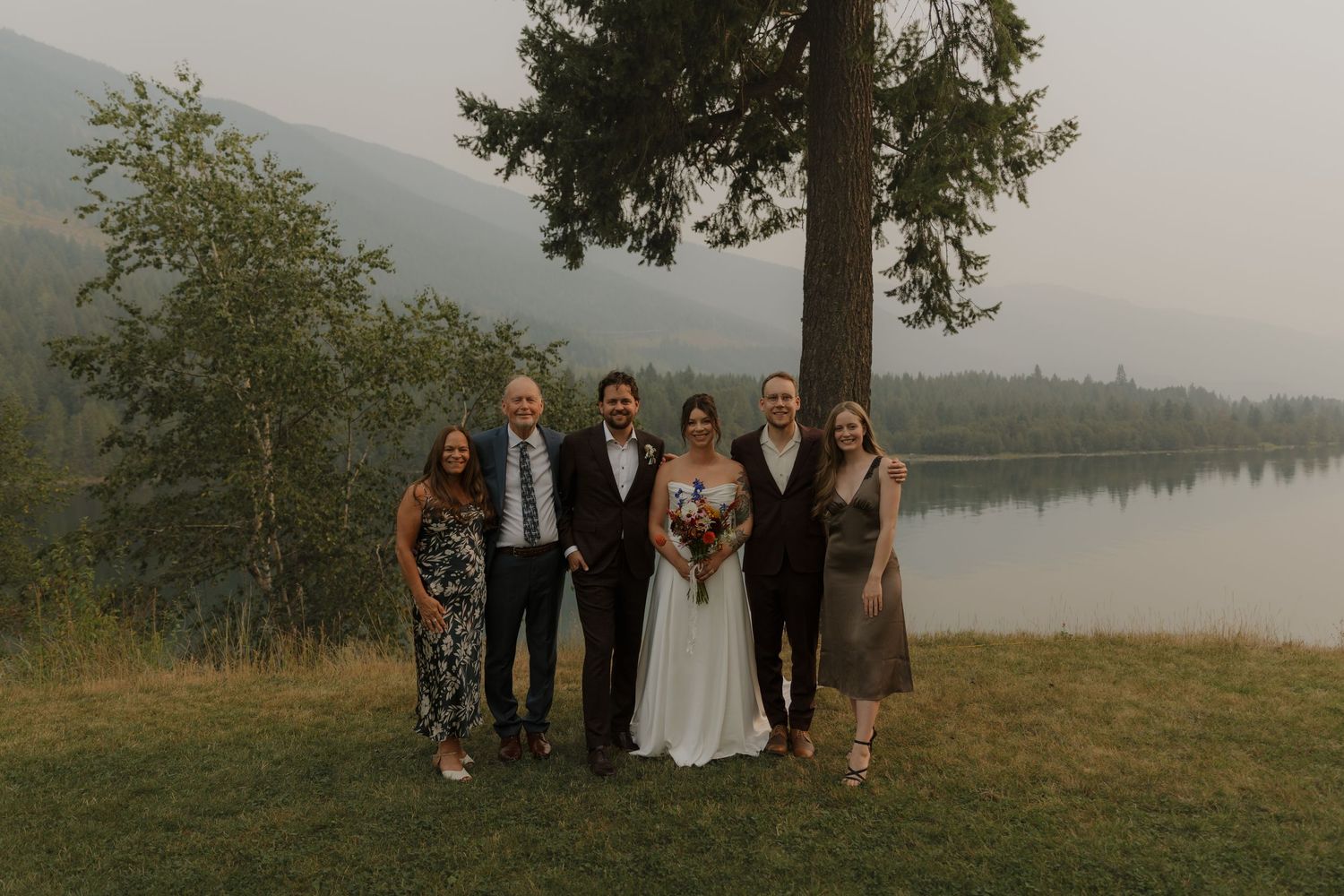 Wedding party group portrait standing by a serene misty lake under a tall pine tree.