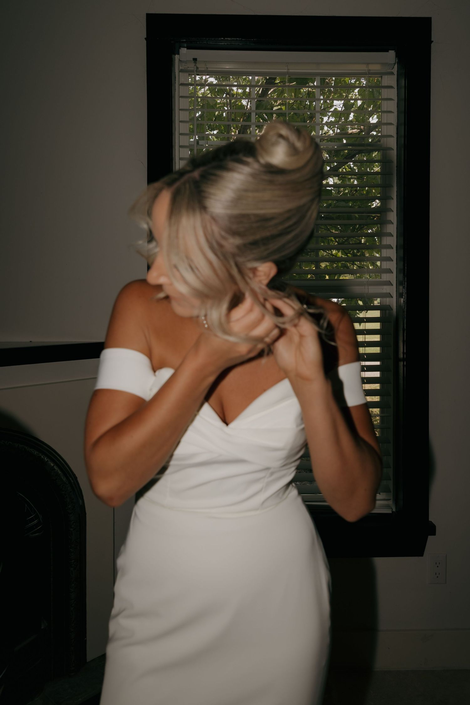 Bride in off-shoulder white wedding gown adjusting hair updo near window with blinds.