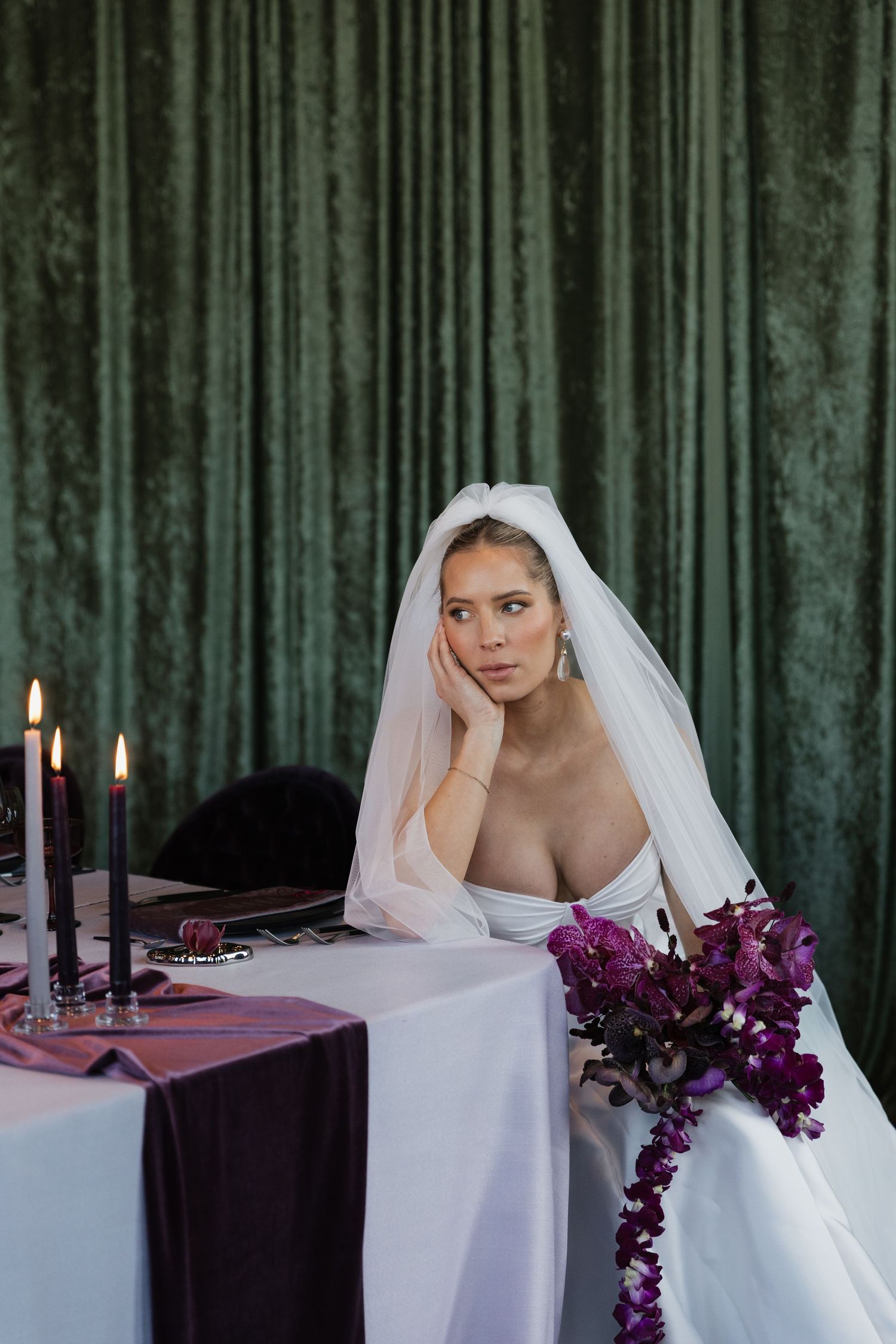 Elegant bride in white gown and veil sits at decorated wedding table with purple flowers and dark candles.