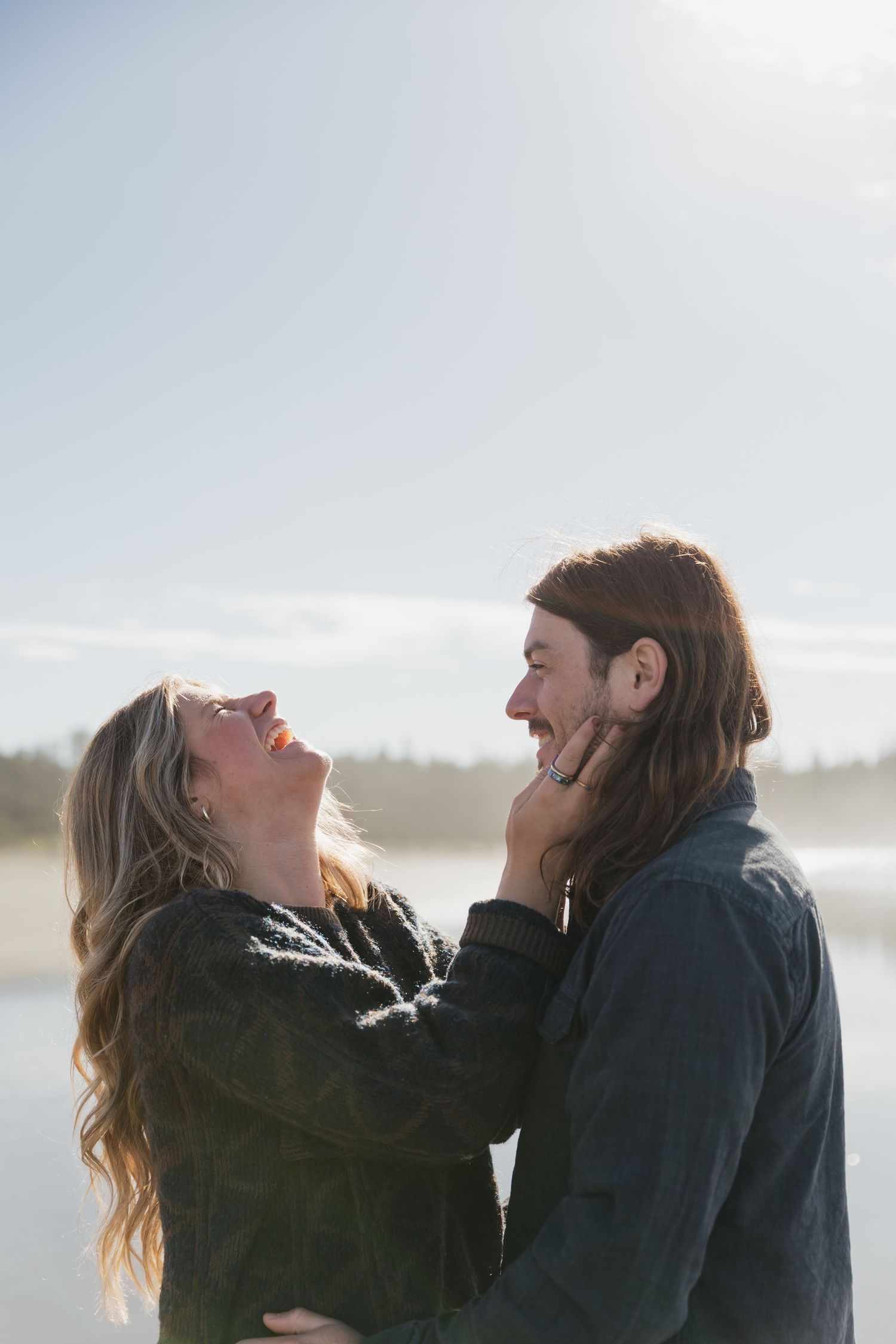 A joyful couple laughing and embracing on a sunny beach, the woman holding the man's face tenderly.