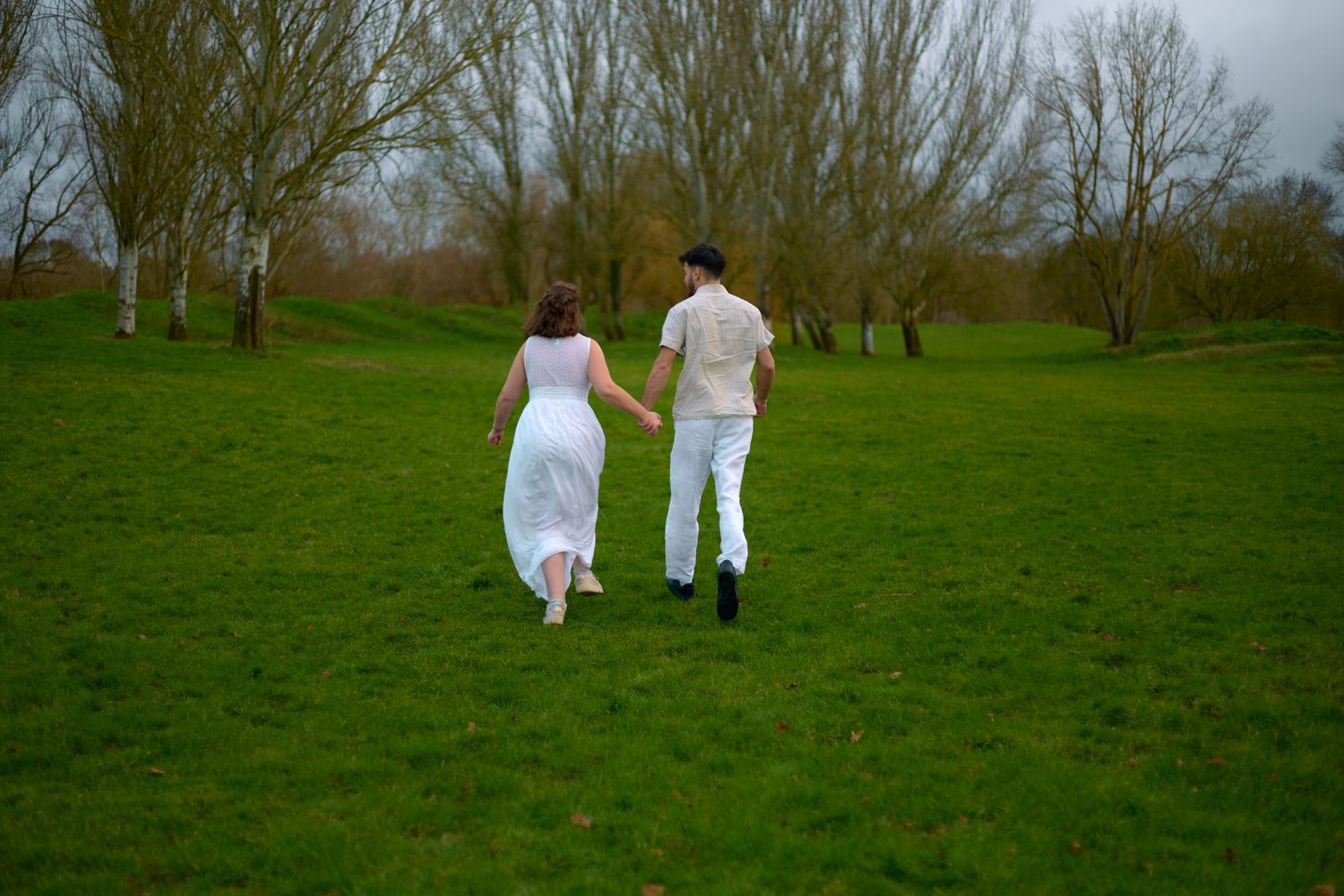 A couple in white clothing walks hand in hand across a green field with bare trees.