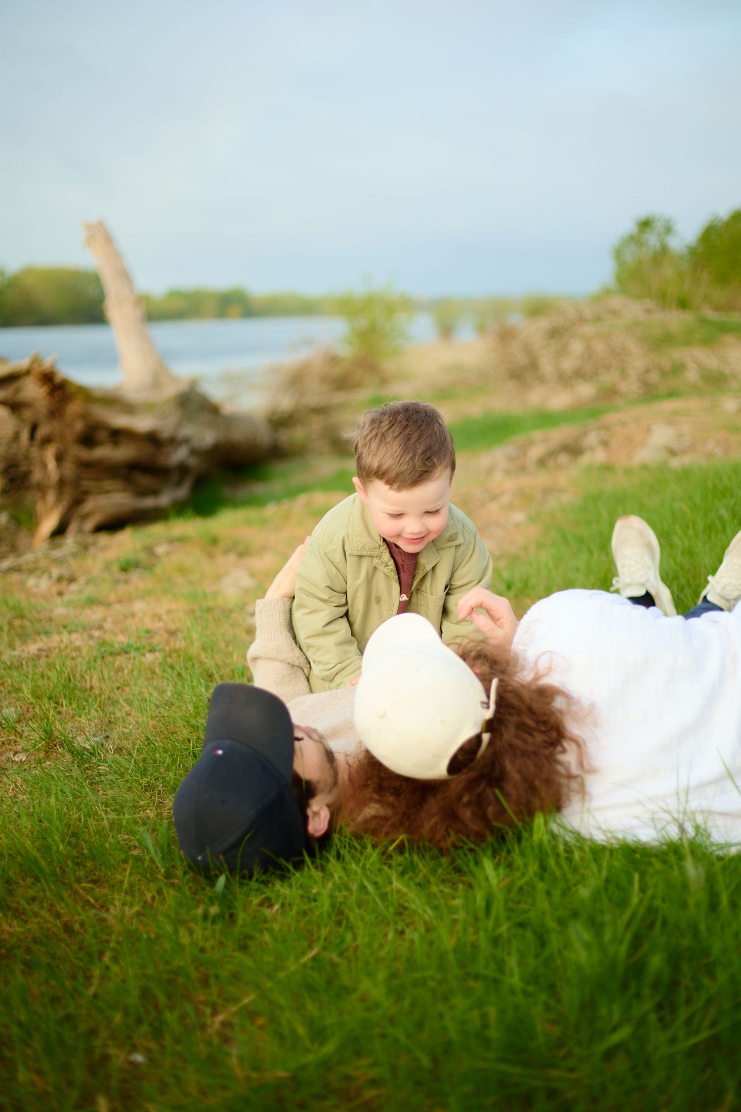 A joyful young boy plays with two adults lying on green grass near a river on a cloudy day.
