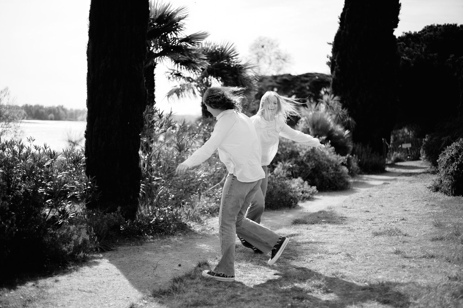 Black and white photo of two people dancing joyfully on a garden path lined with palm trees and lush shrubs.