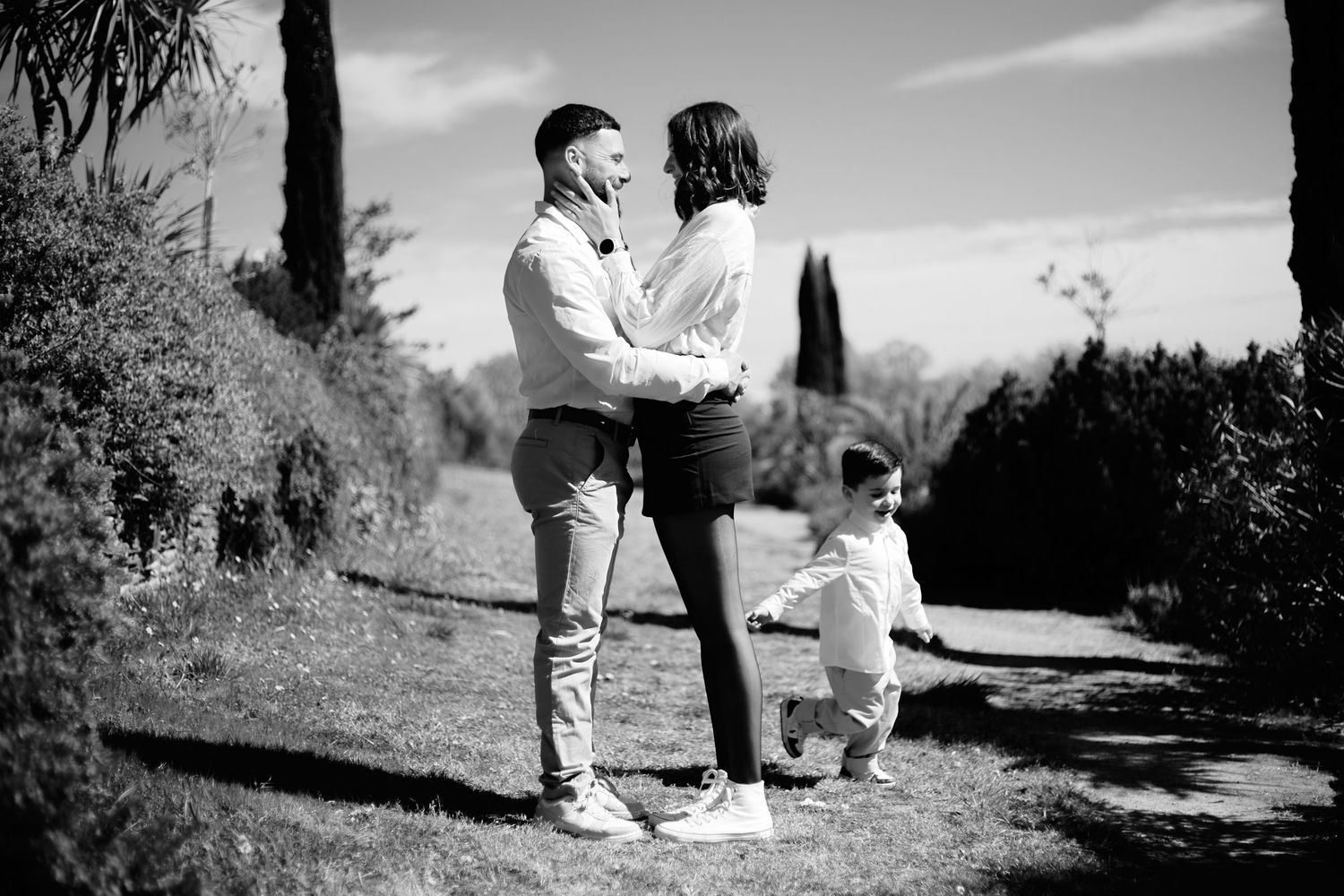 Black and white photo of a couple embracing outdoors while a toddler plays nearby in a garden setting.