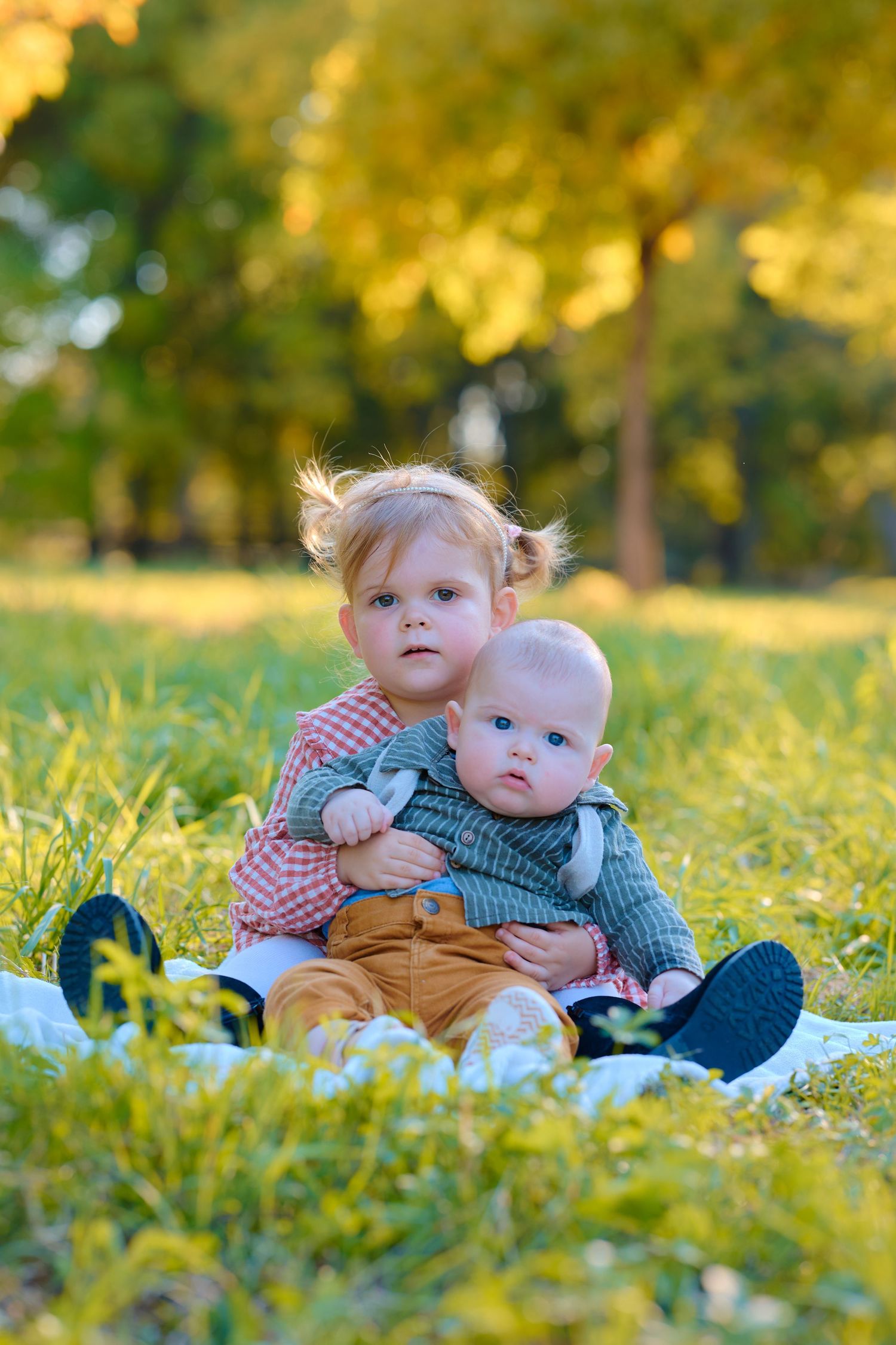 Two young siblings sitting on grass in a sunny autumn park, older toddler girl hugging baby brother.