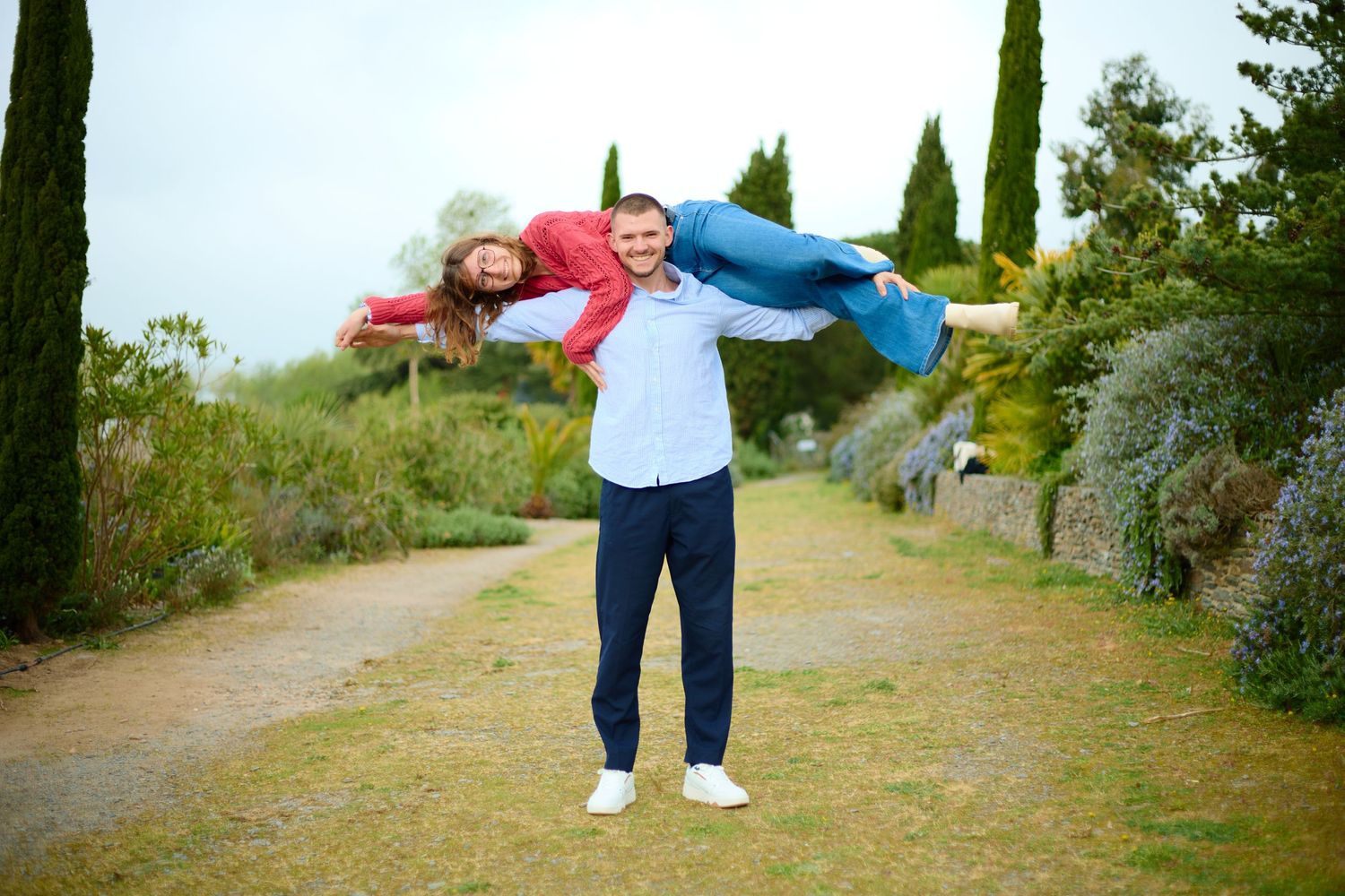 Man playfully lifting woman in red shirt outdoors, both smiling in a lush garden pathway with cypress trees.