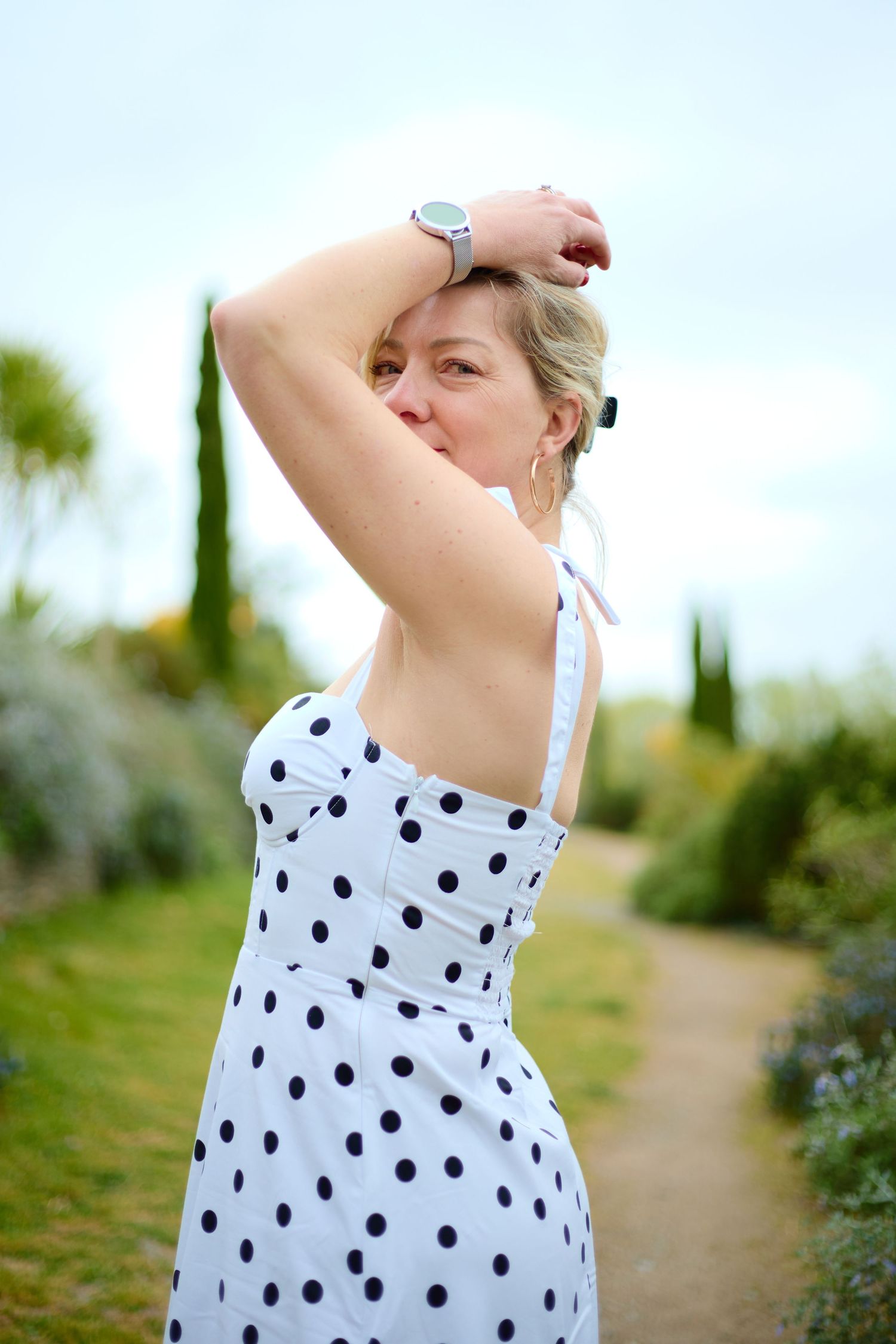 Woman in white polka dot sundress posing in a garden path with arms raised, surrounded by green trees.
