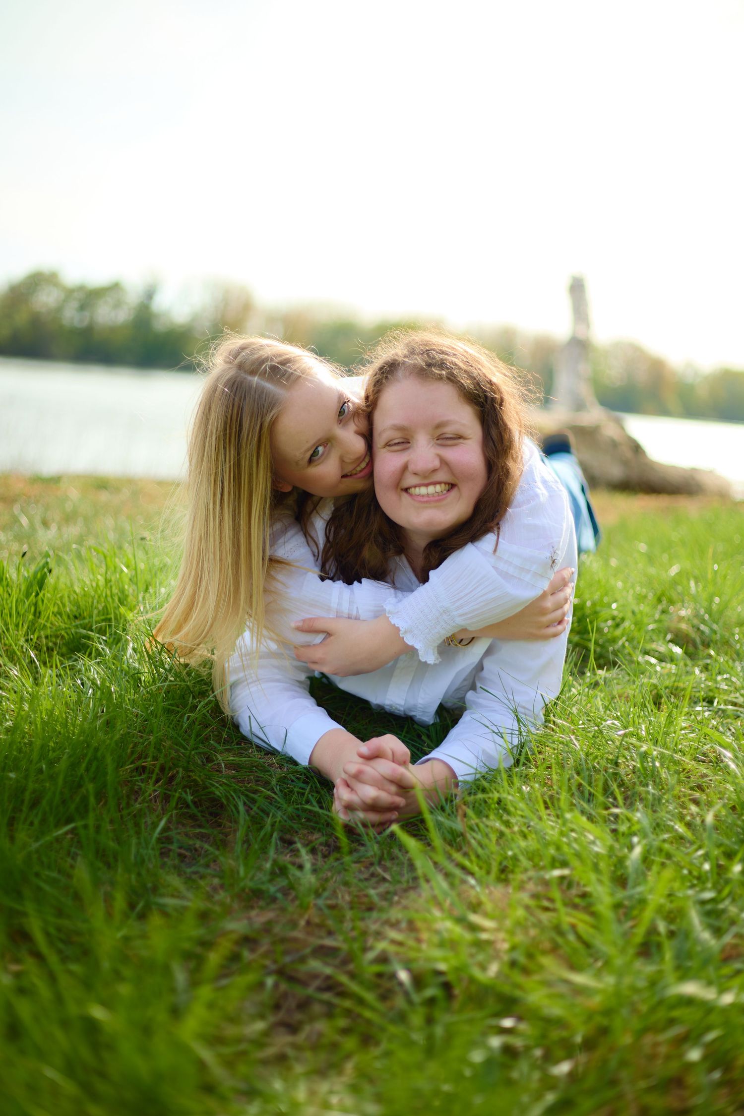 Two young women laughing and hugging on green grass near a lake on a sunny day.