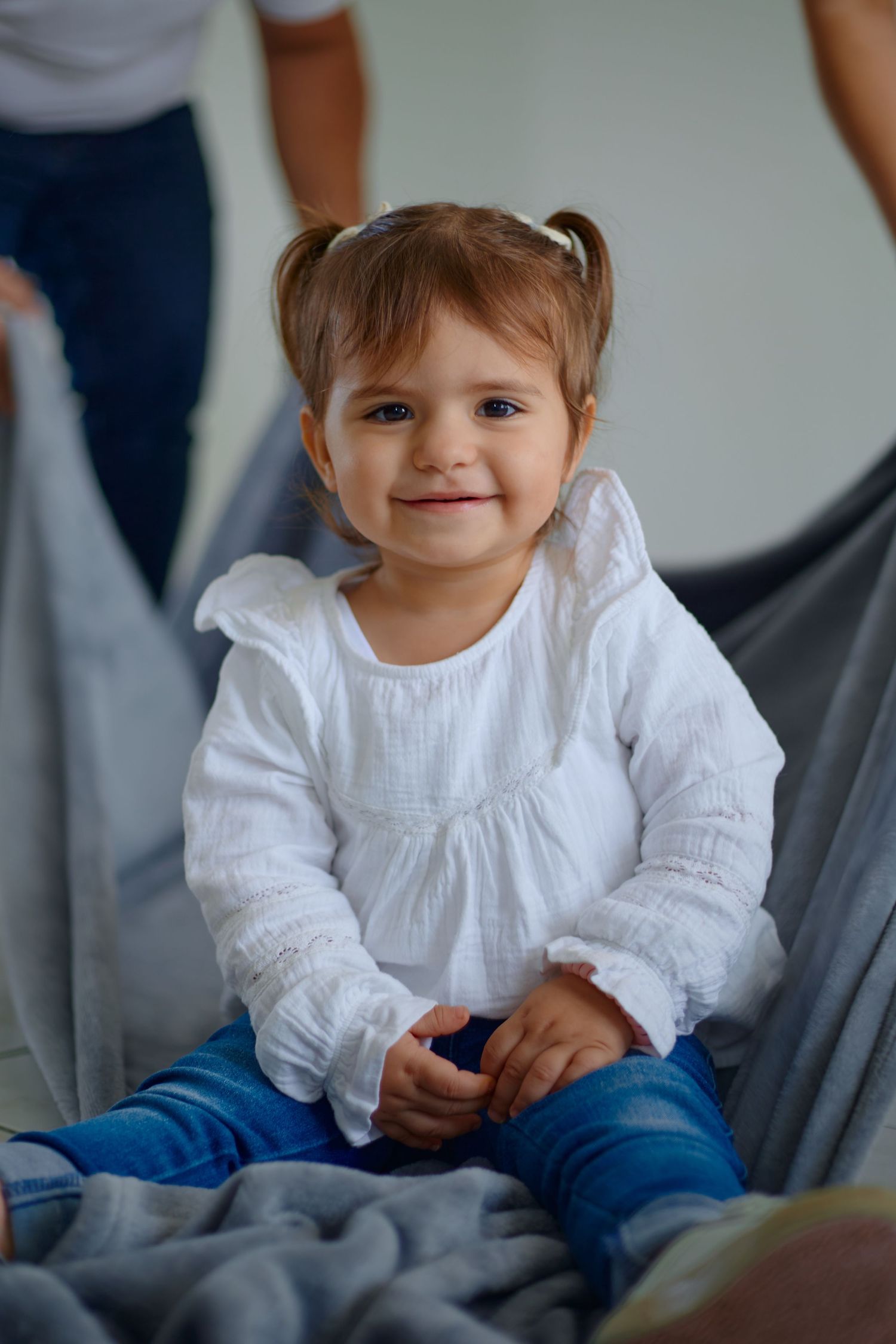 A toddler in a white dress smiles while playing on grey furniture indoors.