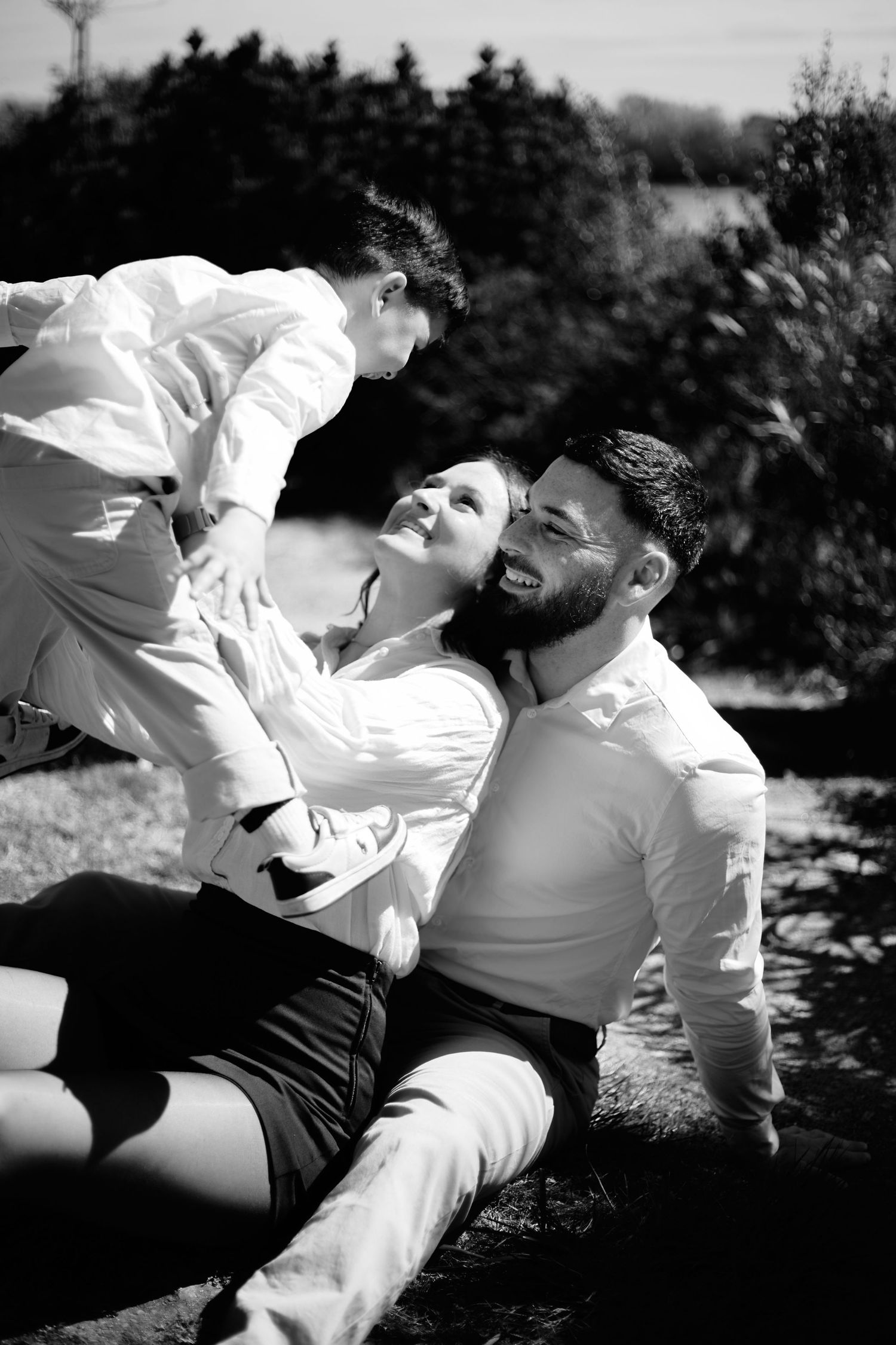 Black and white photo of a happy family with parents sitting outdoors, joyfully lifting their young child in the sun.