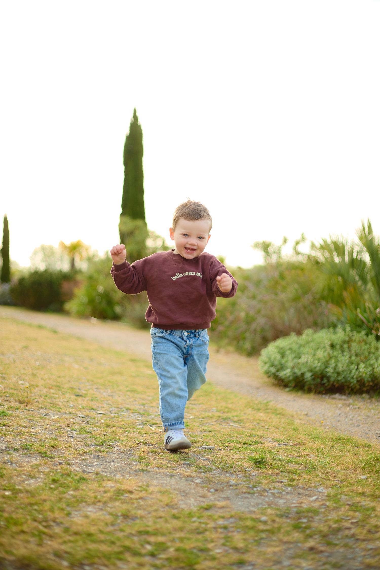 A happy toddler boy in a maroon sweatshirt and jeans runs along a garden path with lush greenery.