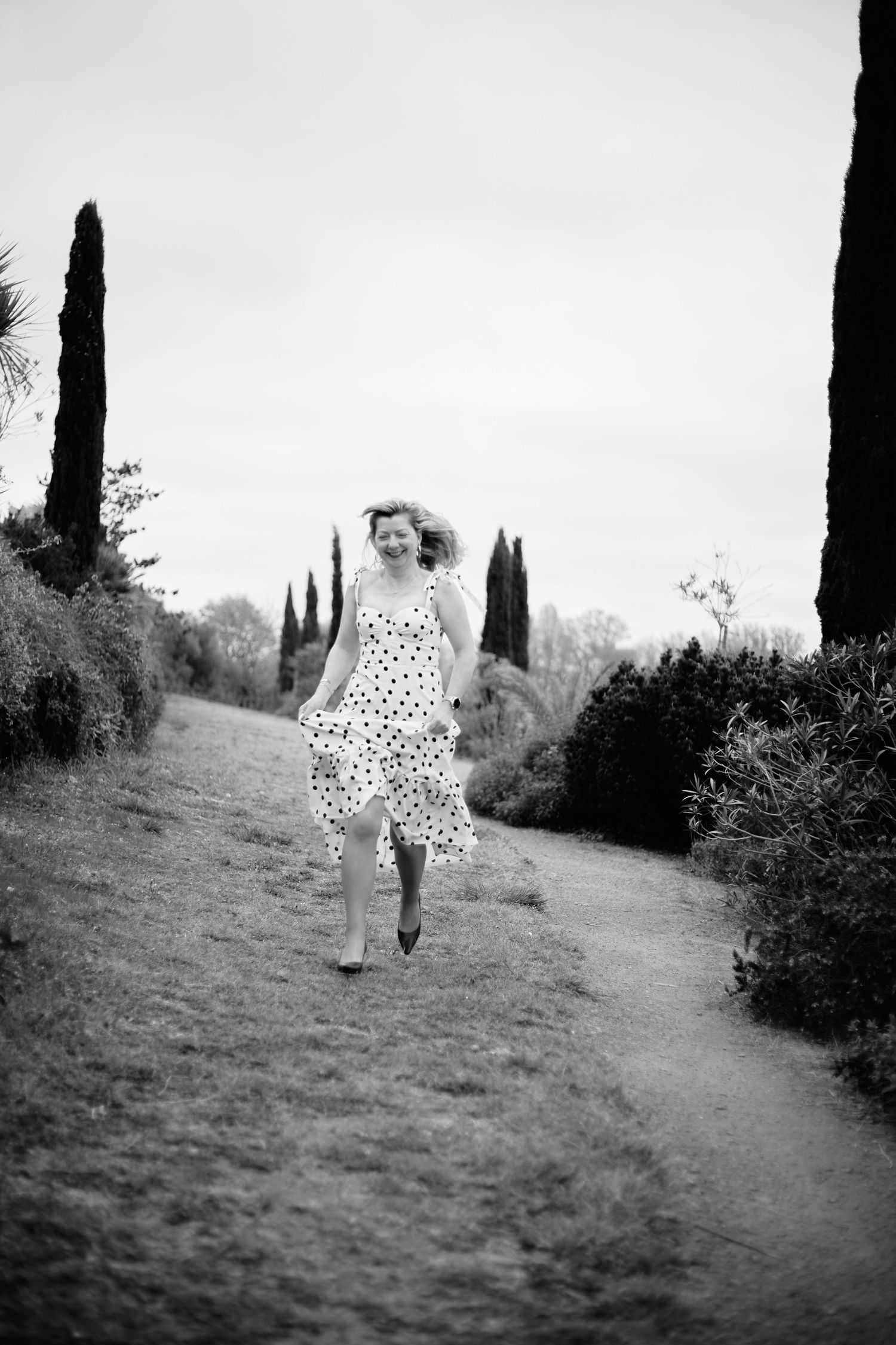 Woman in polka dot dress running joyfully down a garden path lined with cypress trees, black and white photo.