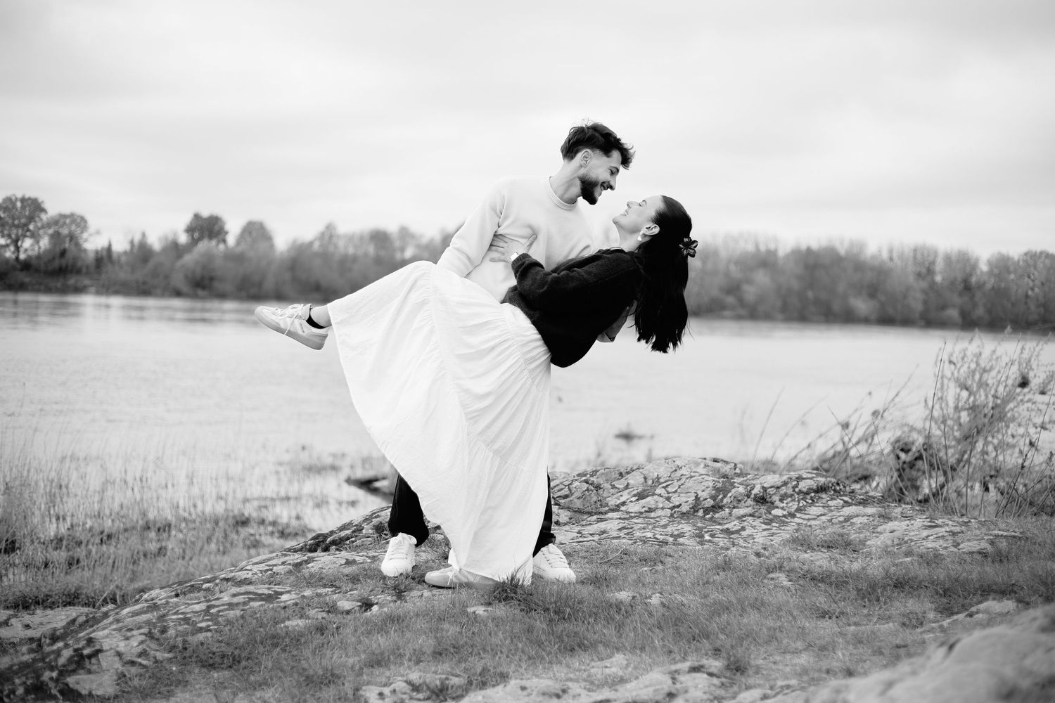 Black and white photo of a couple sharing a romantic dip kiss by a serene lakeside in wedding attire.