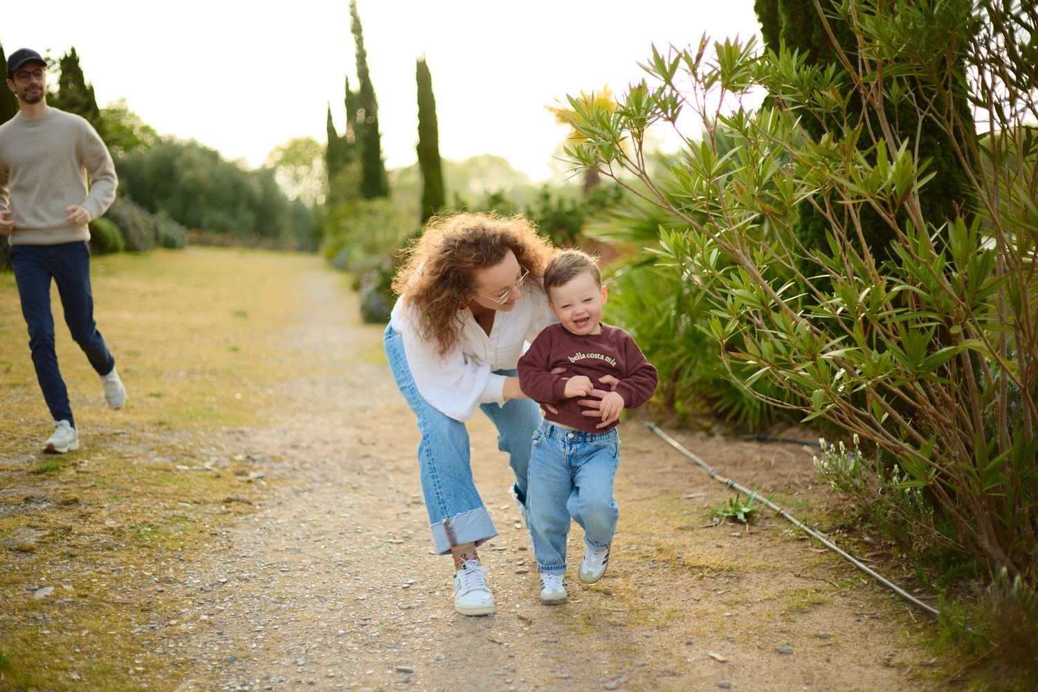 A mother helps her toddler walk on a garden path while a man jogs behind them outdoors.