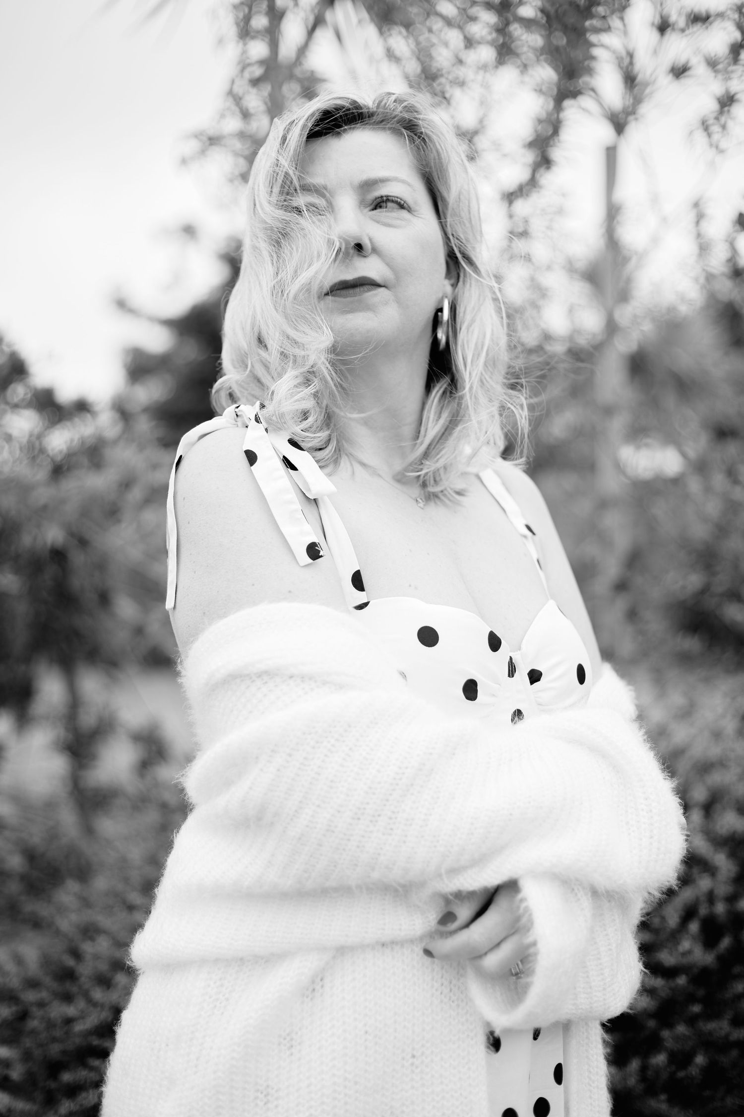 Black and white portrait of a blonde woman in polka dot dress and white knit wrap, standing outdoors.