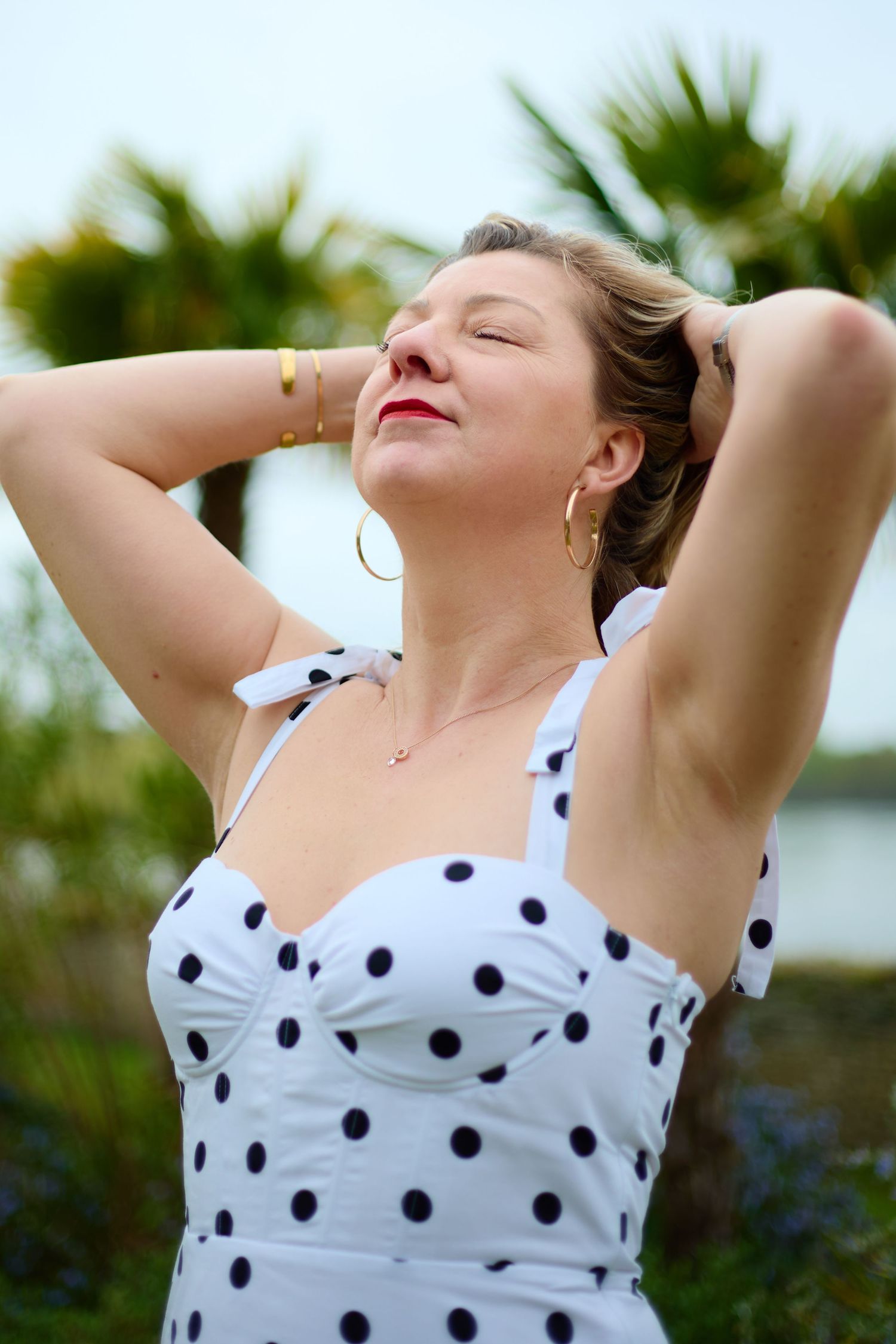 Woman in white polka dot dress with hands behind head, eyes closed, enjoying sunshine near palm trees.