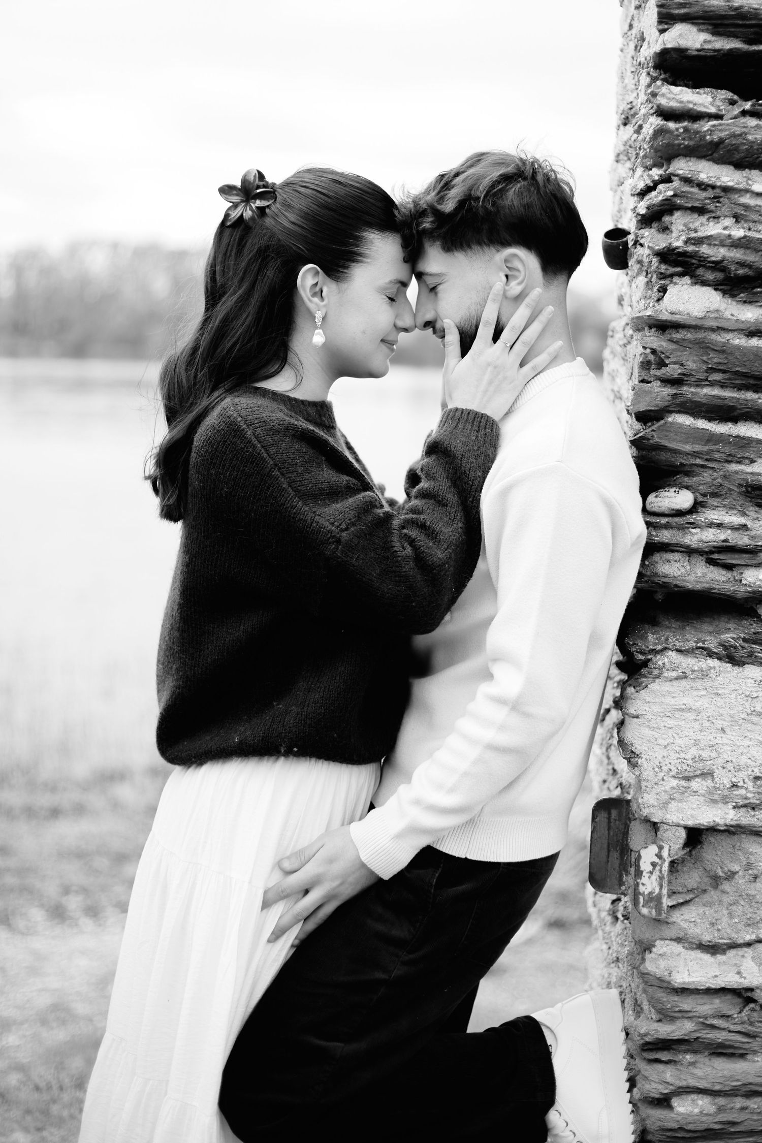Black and white photo of a romantic couple leaning against a stone pillar, touching foreheads tenderly outdoors.